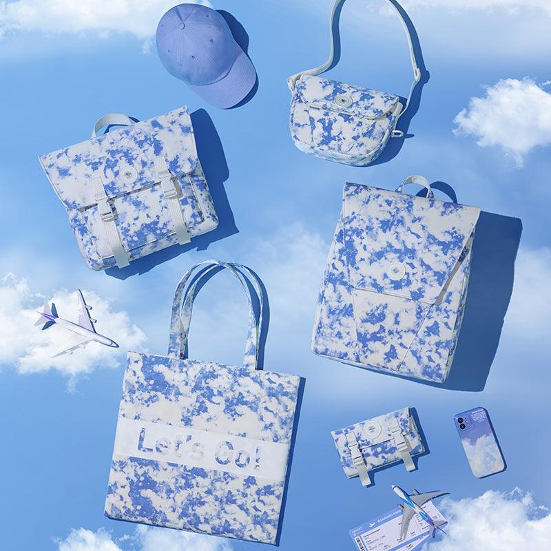 Blue cloud-print backpacks, tote, crossbody bag, cap, and phone case against sky with planes