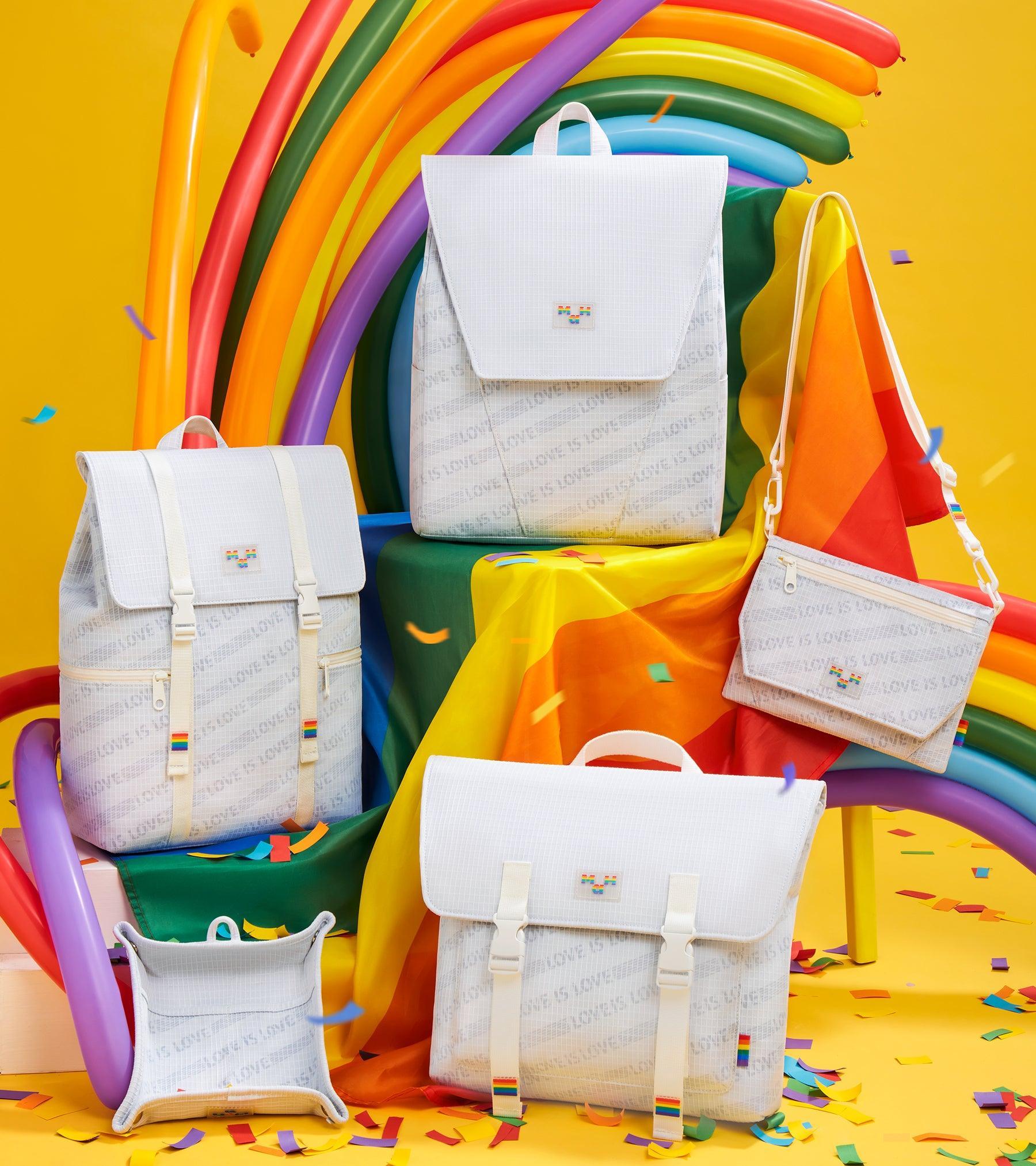 White rainbow-themed backpacks and bags with 'Love is Love' print, displayed with colorful balloons and confetti