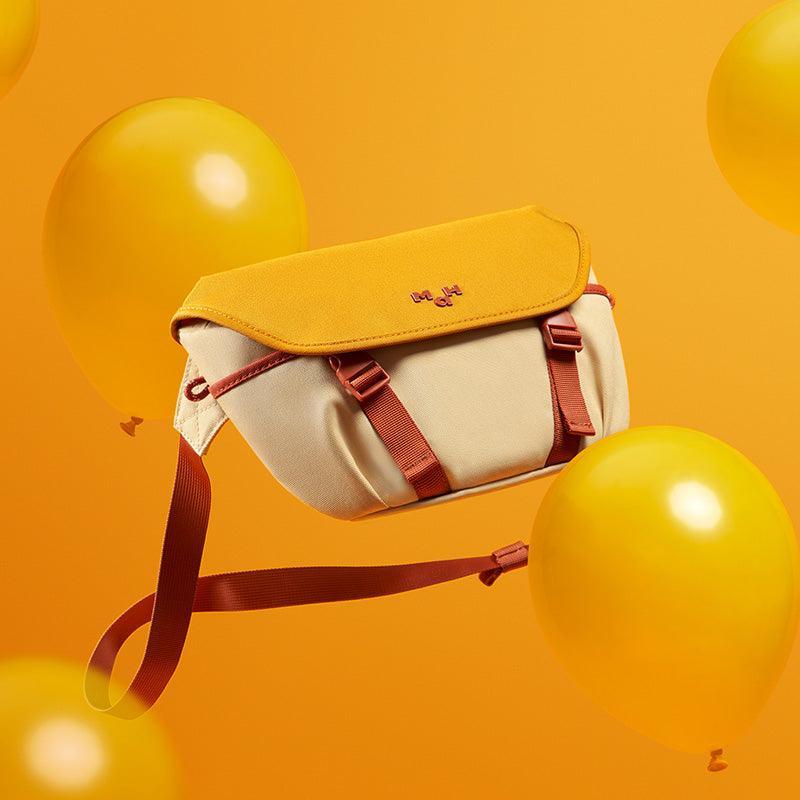 Stylish yellow and cream crossbody bag with red straps, floating among yellow balloons on orange background