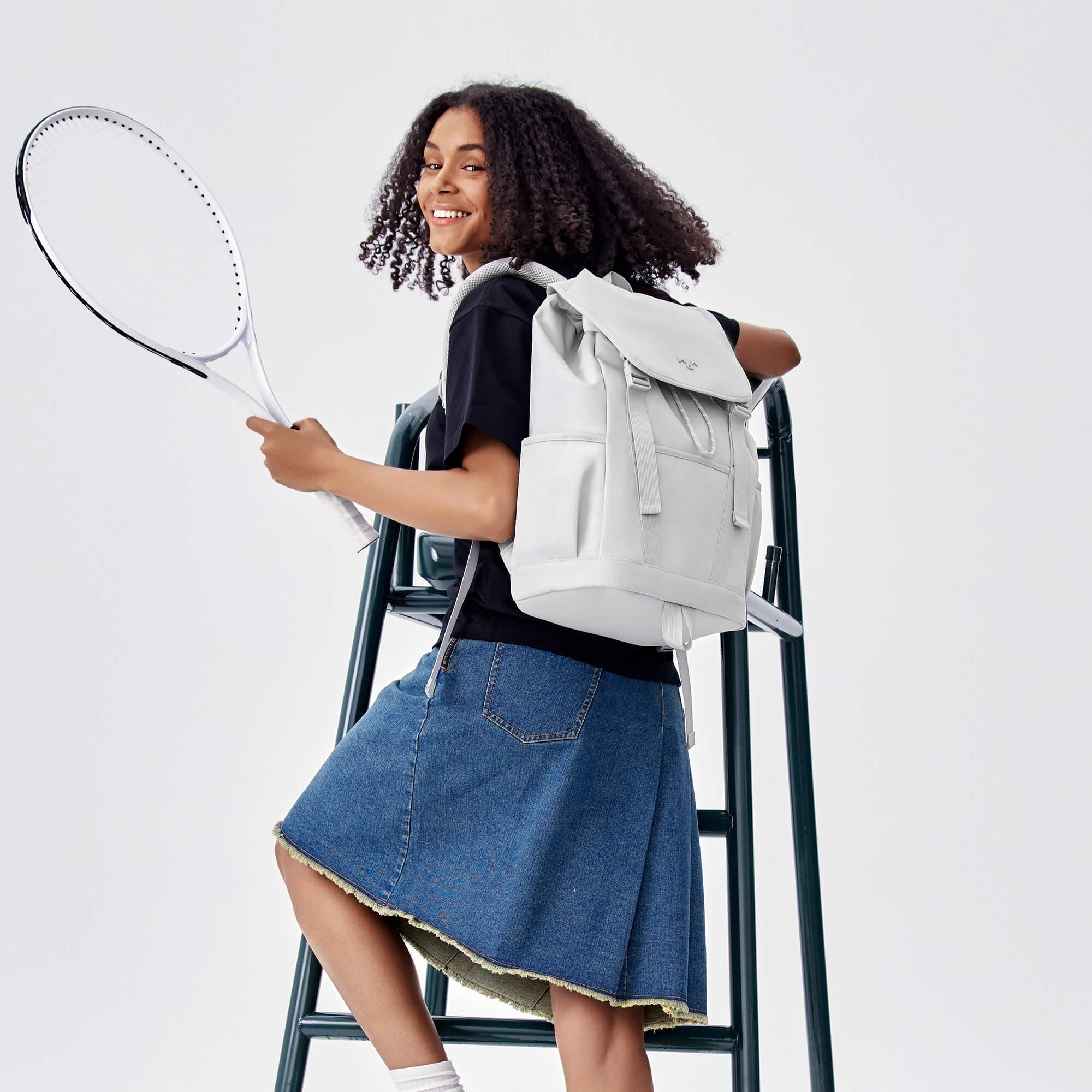 Woman with curly hair wearing light gray backpack, holding tennis racket, seated on chair
