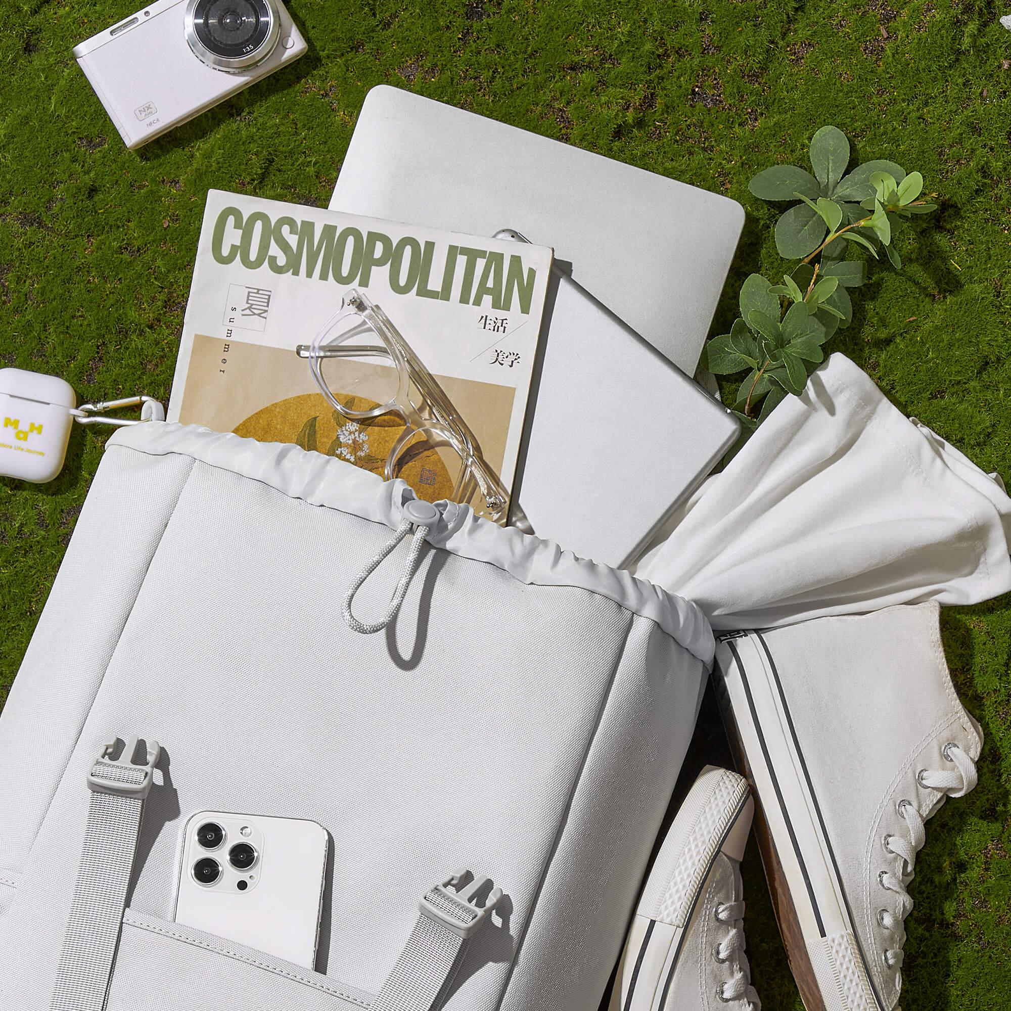 White drawstring backpack with phone, laptop, magazine, and sneakers on grass — stylish everyday carry essentials