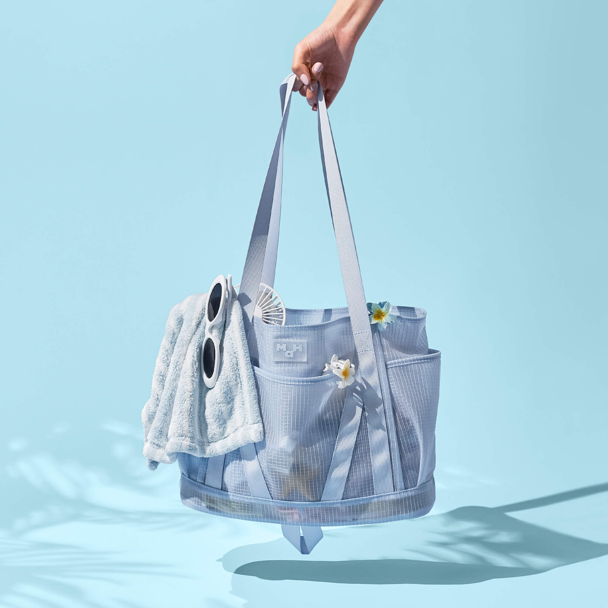 Light blue mesh beach tote bag with sunglasses, towel, and flowers, perfect for summer outings and poolside essentials.