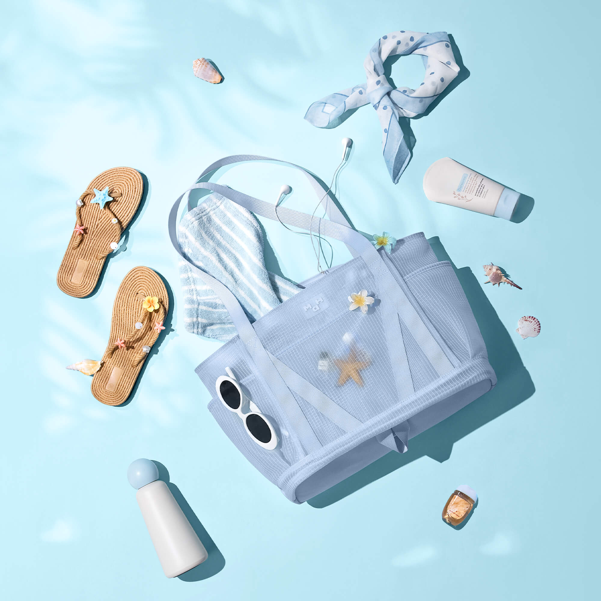 Light blue beach tote bag with sunglasses, flip flops, towel, and summer accessories on a pastel background