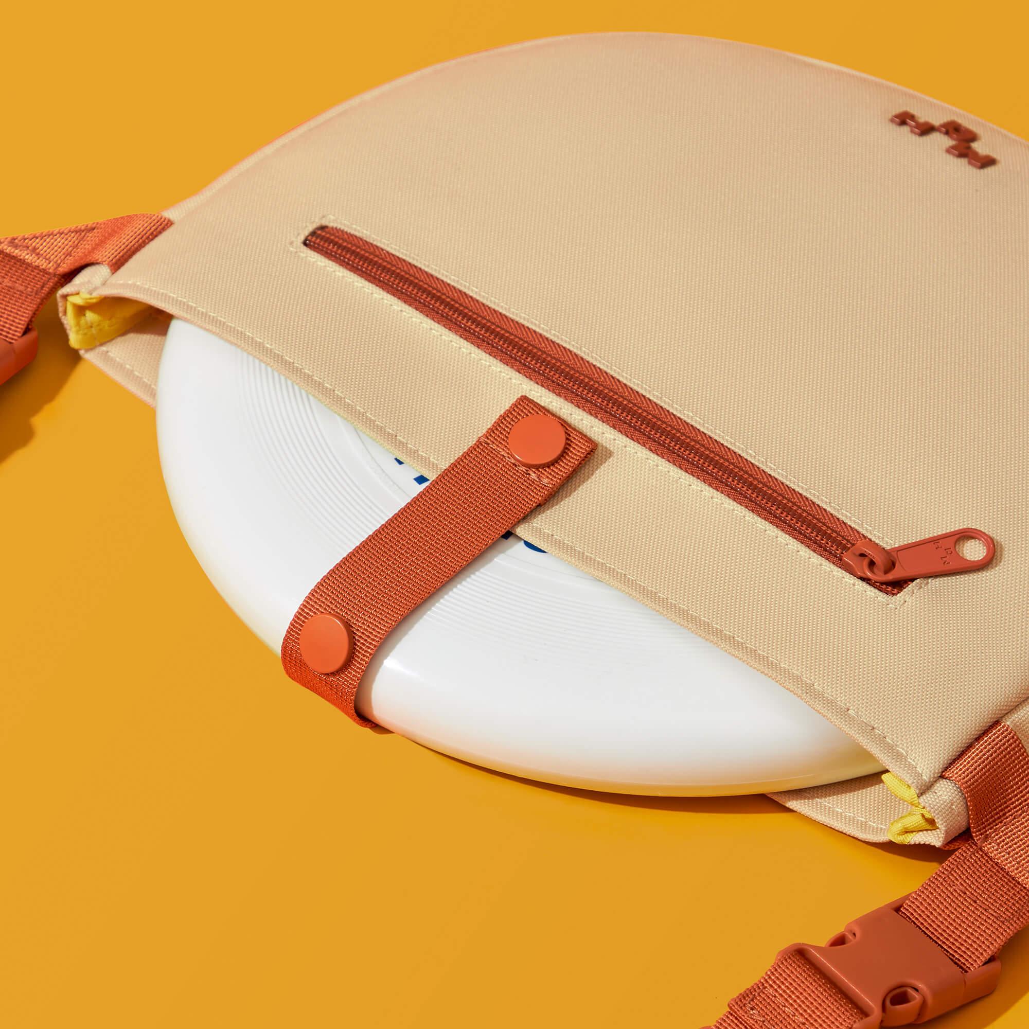 Beige frisbee bag with orange straps and zipper, holding white disc against yellow background