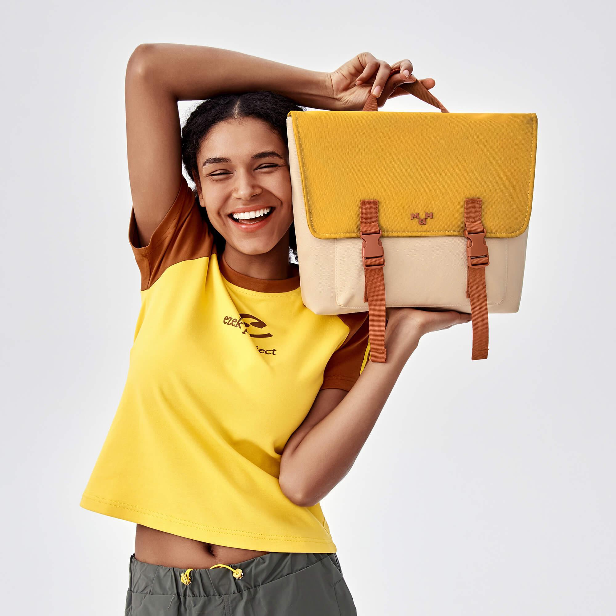 Smiling woman holding yellow and beige satchel bag with brown straps, stylish everyday accessory