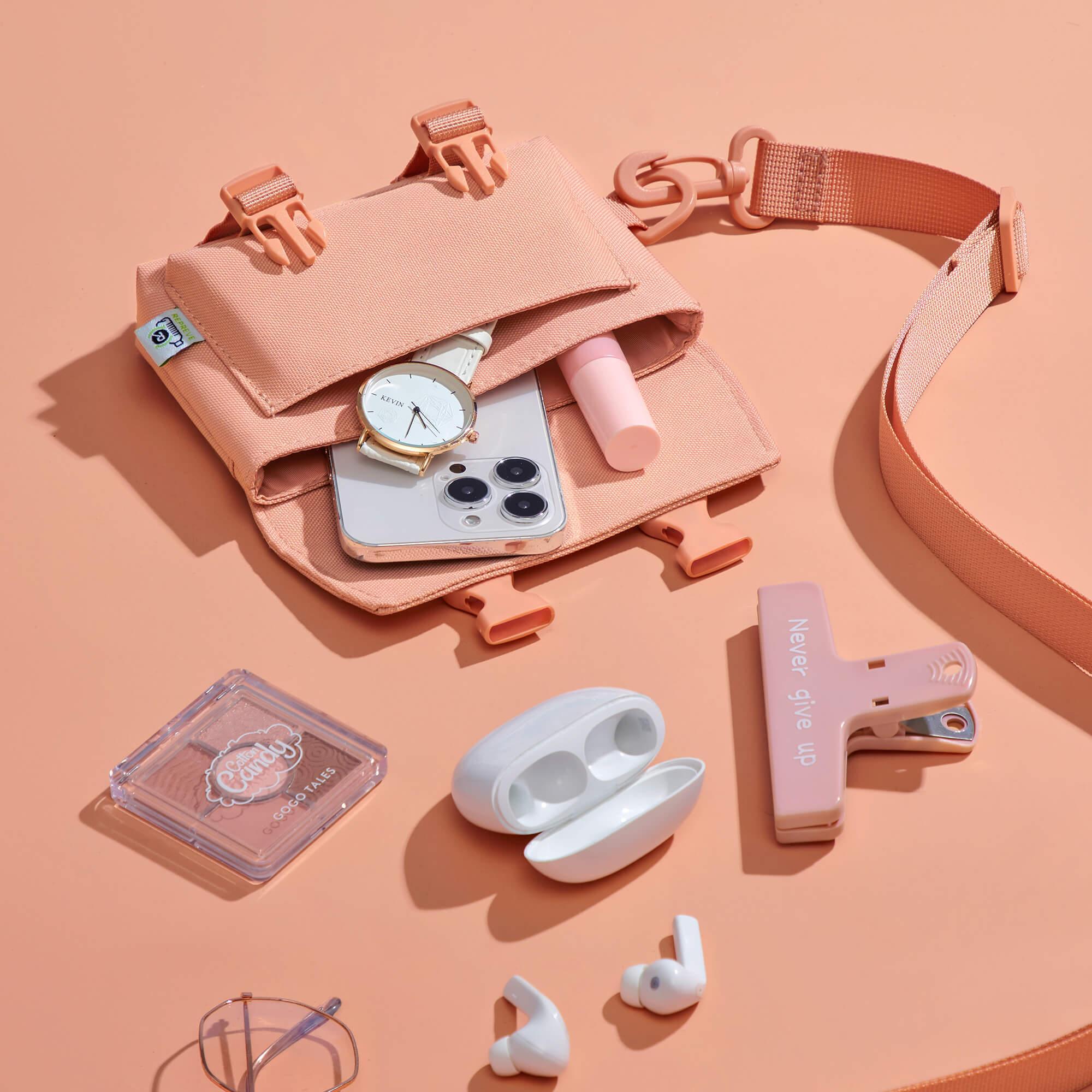 Pink crossbody bag with phone, watch, earbuds, makeup, and accessories on peach background