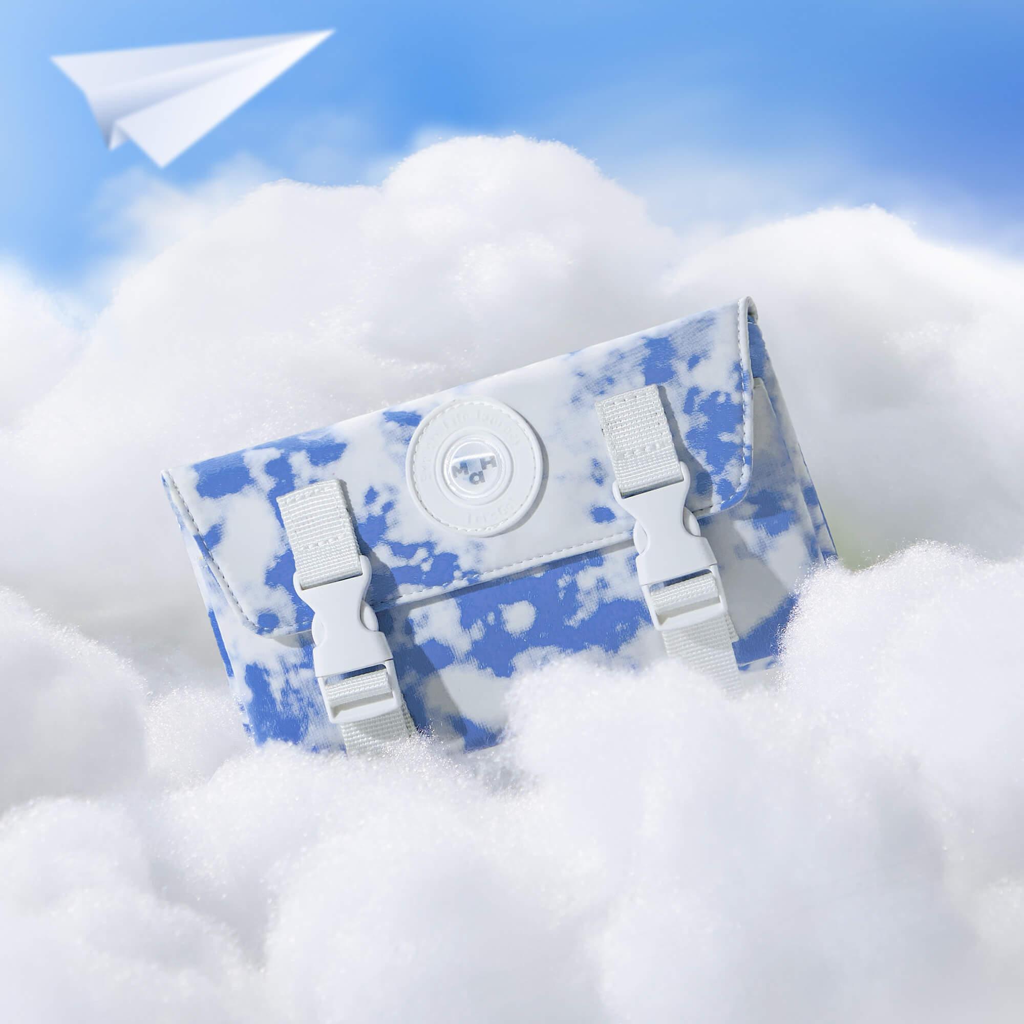 Blue and white cloud-patterned satchel bag with white buckles, nestled in fluffy clouds under a paper airplane.