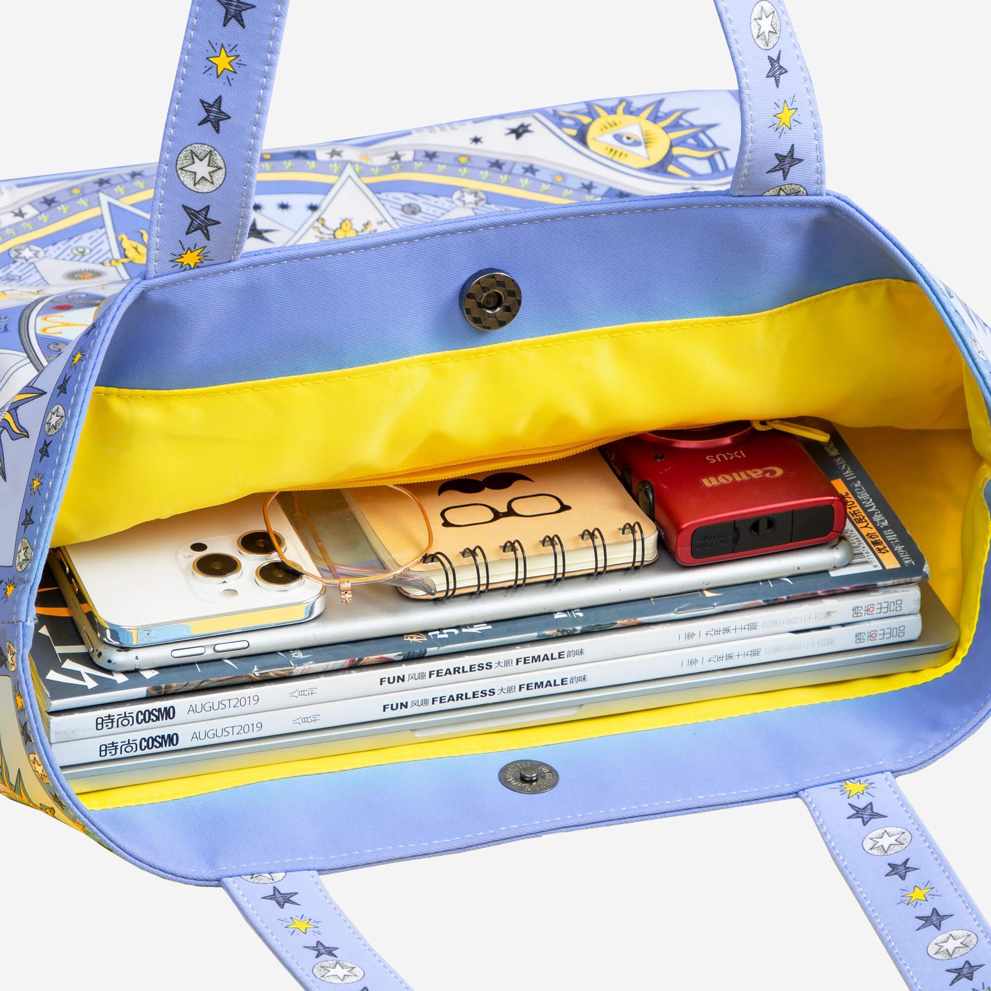 Stylish blue tote bag with star and sun print, yellow lining, holding phone, camera, notebooks, and magazines.