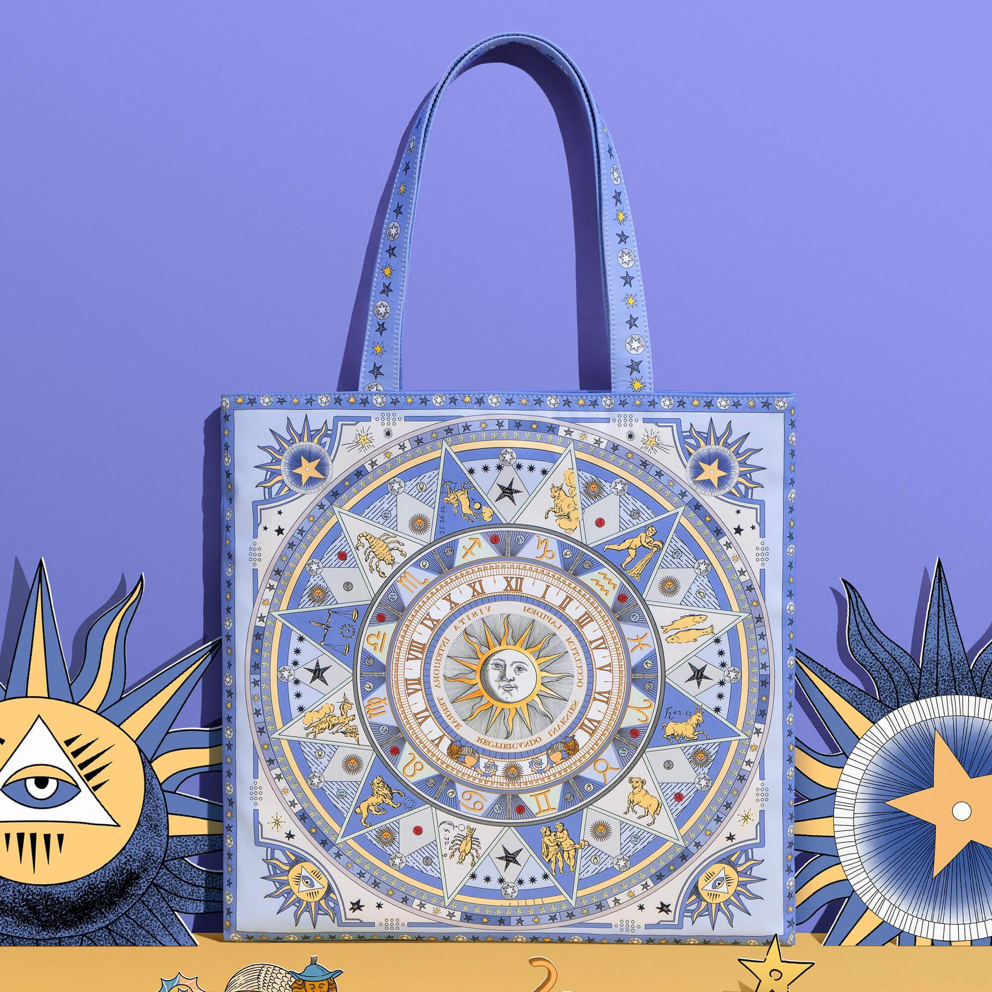 Blue tote bag with astrological zodiac design, celestial symbols, and star patterns on purple background