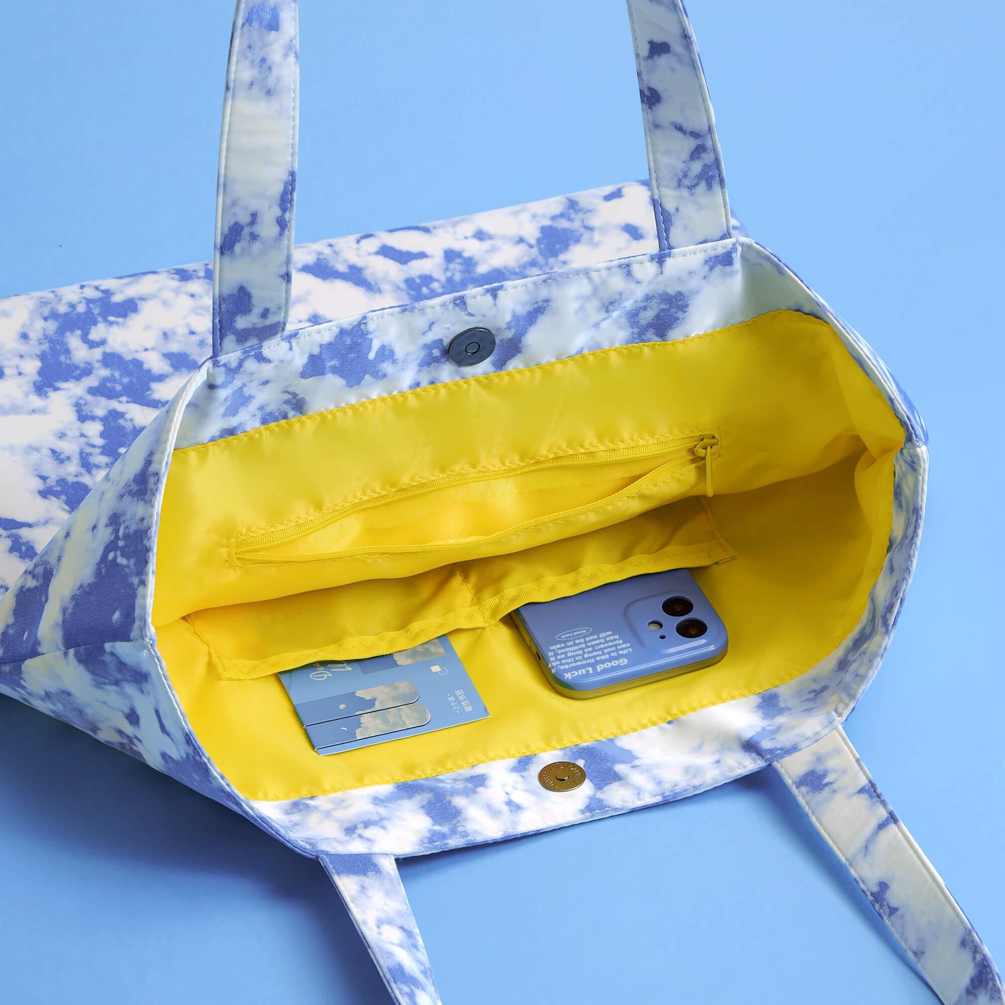 Blue and white cloud print tote bag with yellow interior, holding phone and cards, ideal for daily use.