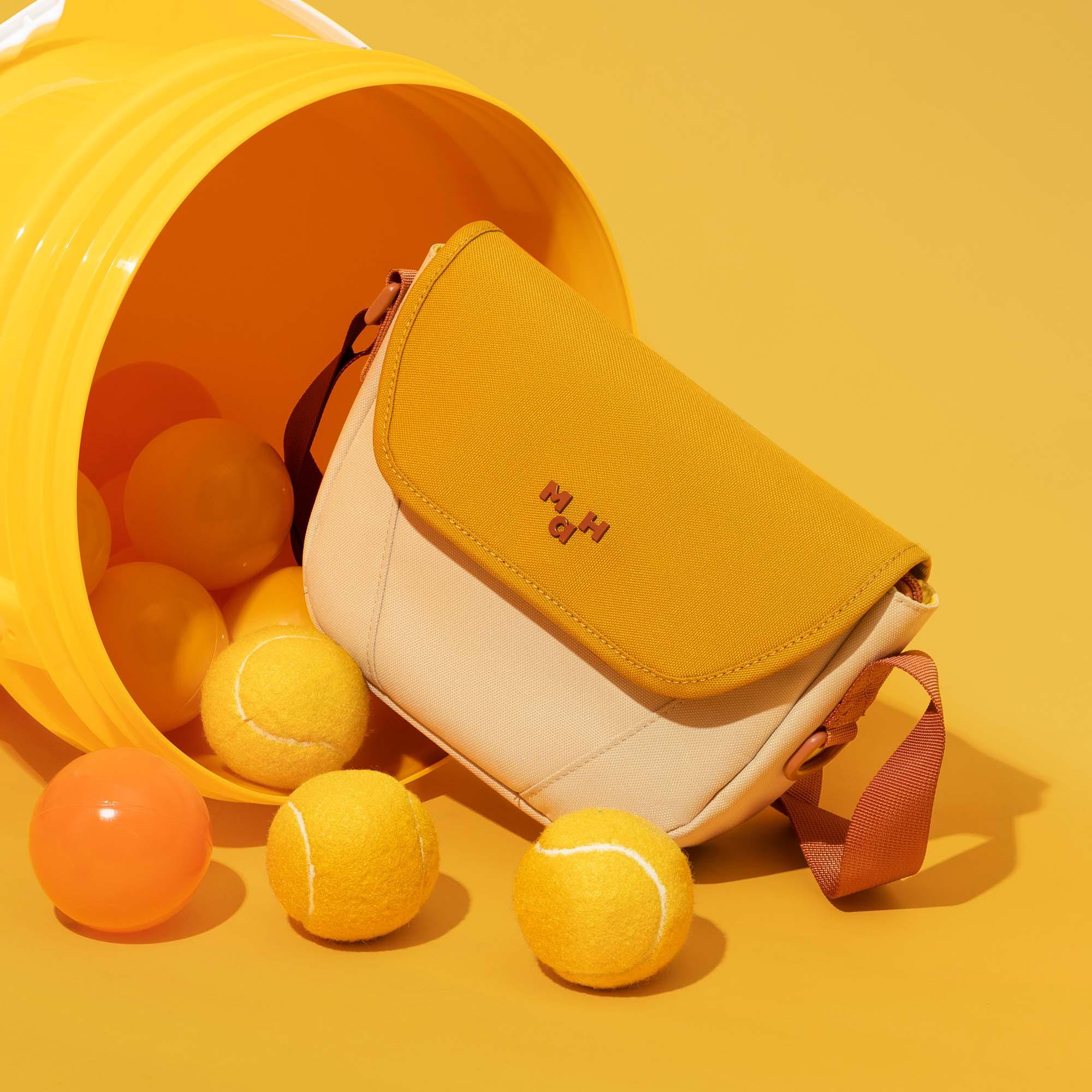 Yellow and cream crossbody bag with tennis balls spilling from a bucket, stylish accessory for active lifestyles