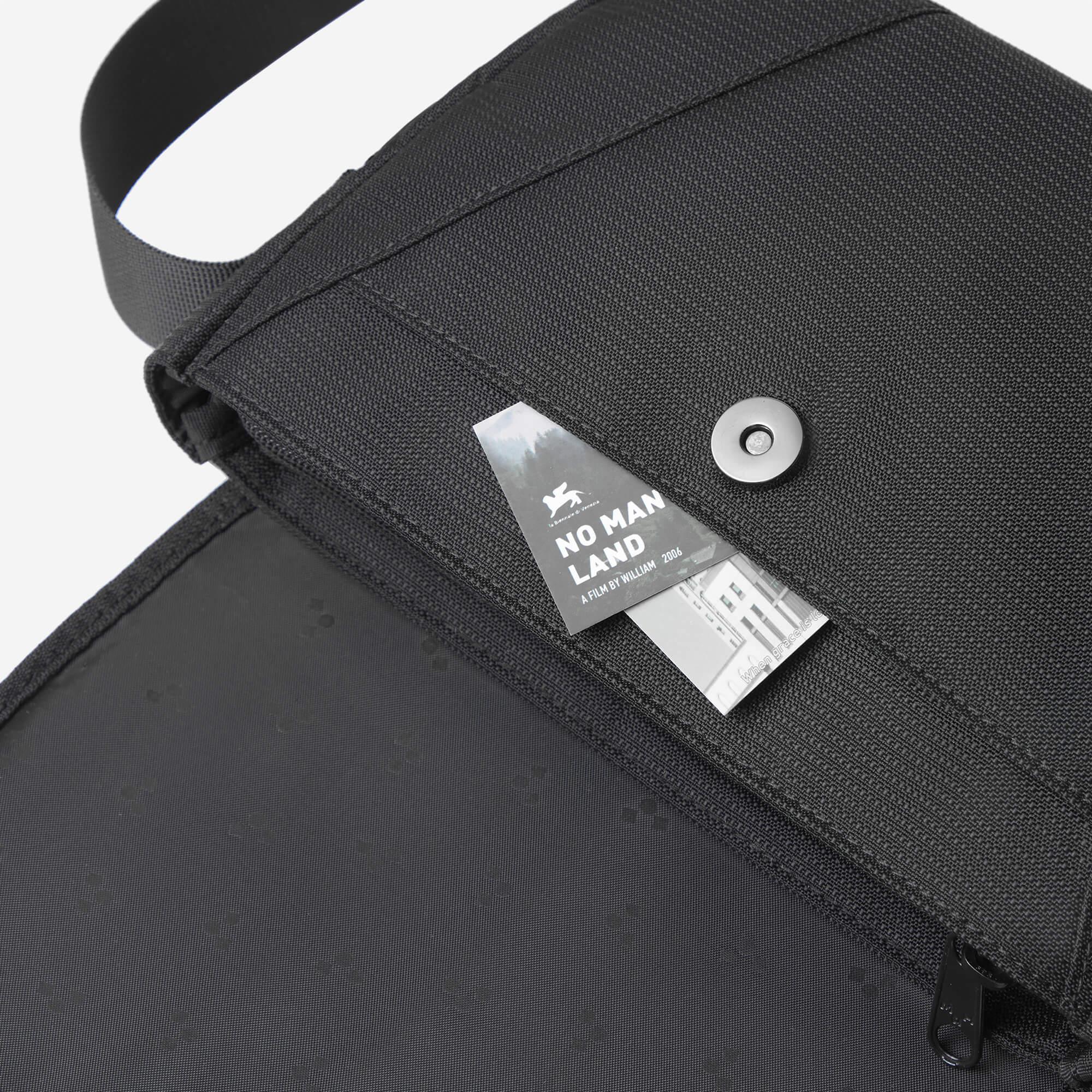 Black textured shoulder bag with 'No Man Land' film tag, metal snap, and zipper detail