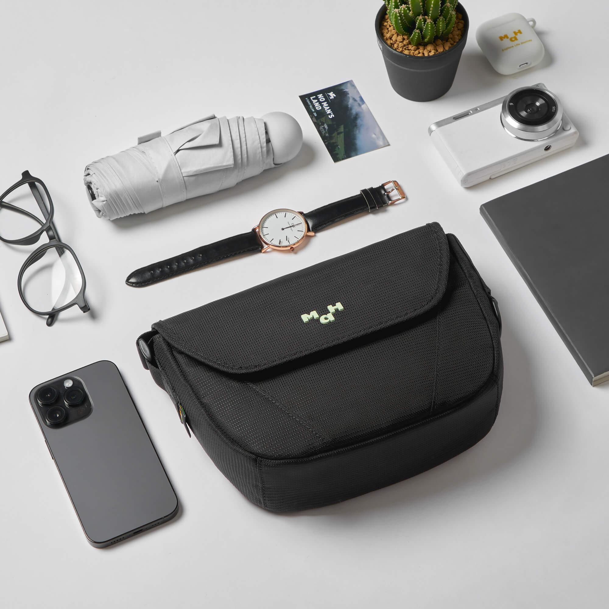 Black MaH crossbody bag with daily essentials: phone, watch, camera, umbrella, and notebook on white desk