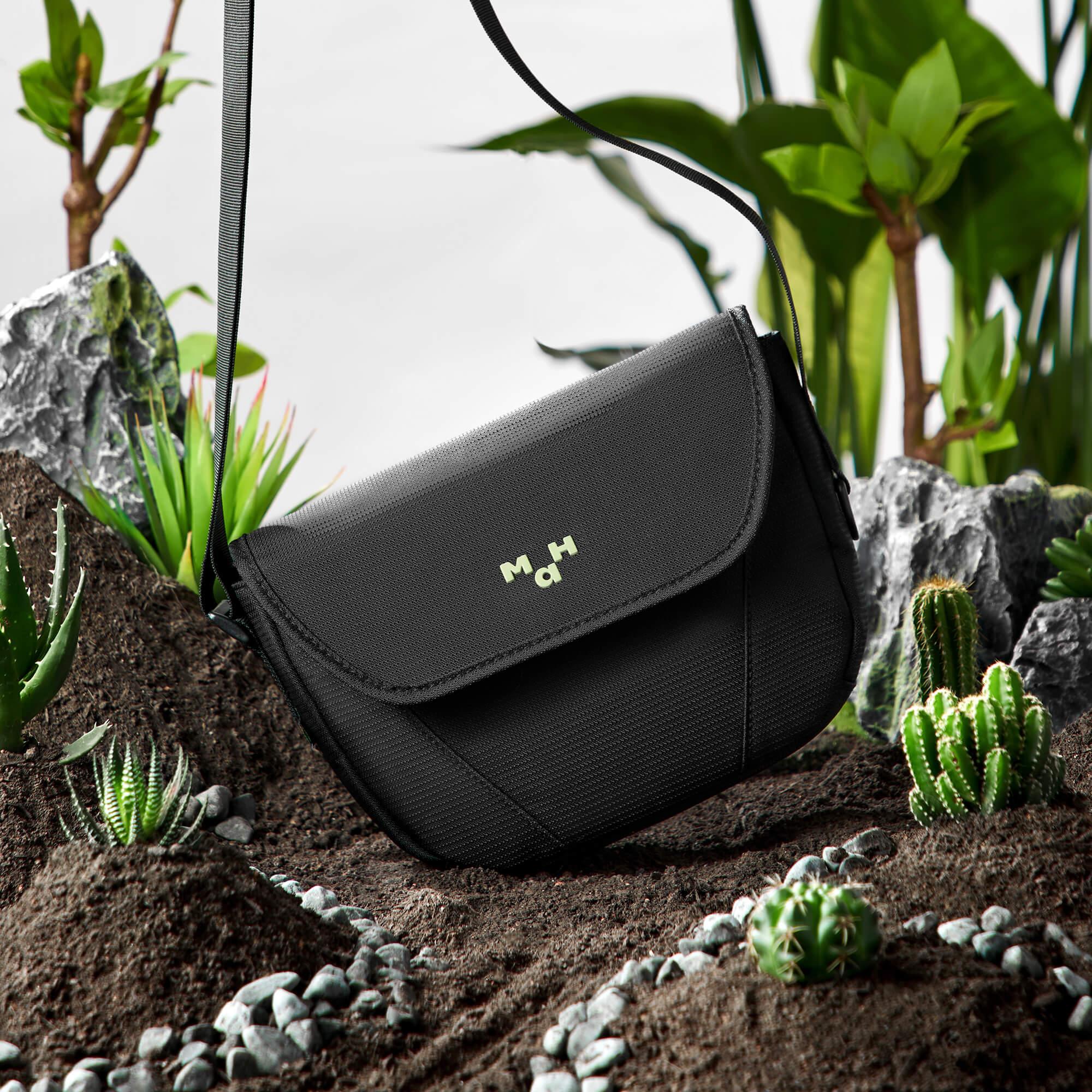 Black crossbody bag with green logo, styled on soil with cacti and succulents for modern lifestyle.