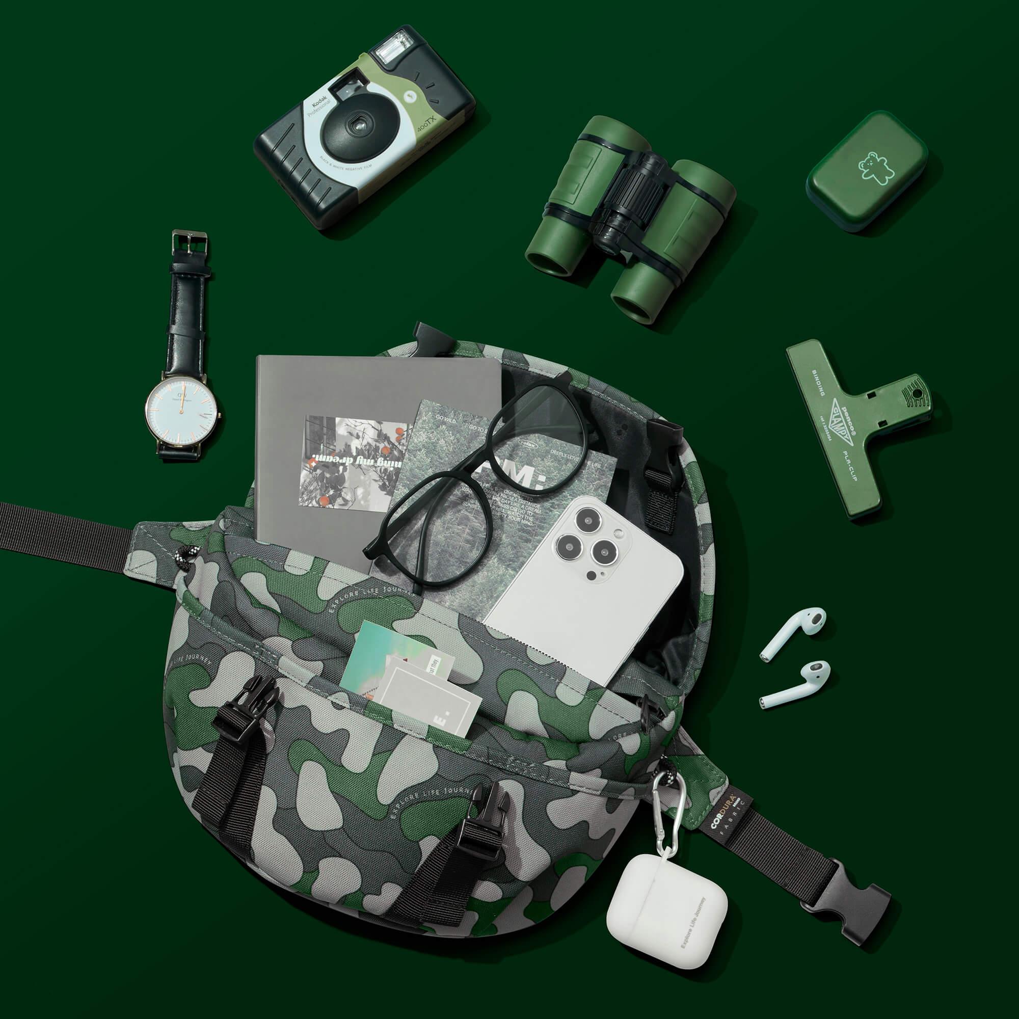Camouflage waist bag with phone, glasses, camera, binoculars, watch, and travel accessories on green background