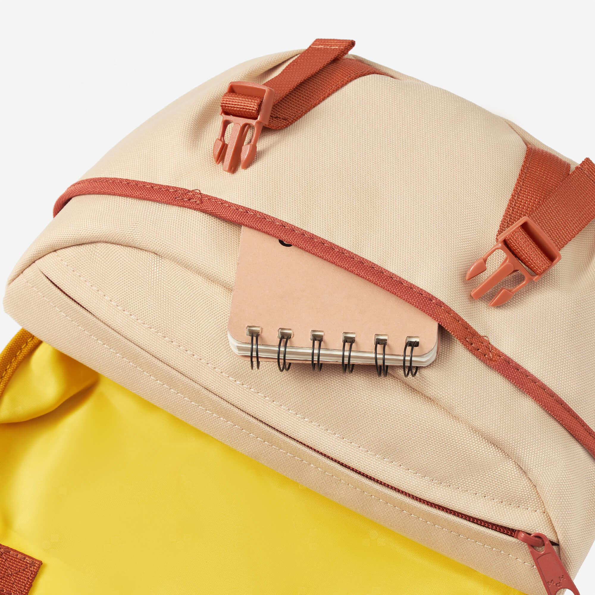 Beige backpack with rust straps, front pocket holding notebook, yellow interior detail