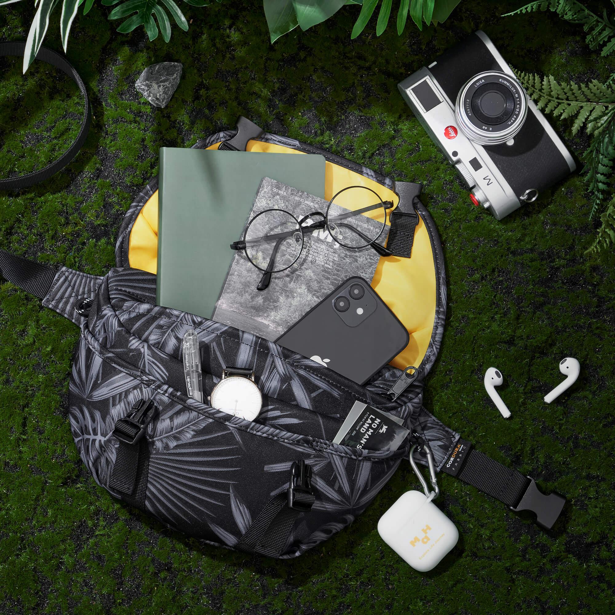 Stylish leaf-patterned waist bag with phone, glasses, notebook, and camera on mossy ground