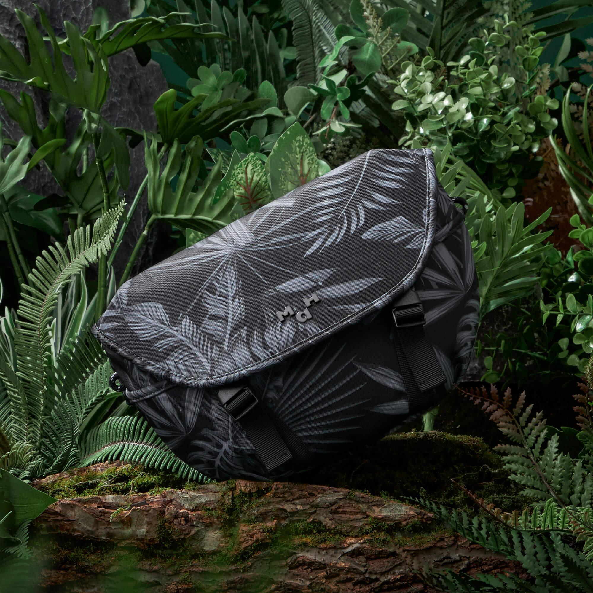 Black tropical leaf print sling bag with adjustable strap, nestled in lush green foliage for outdoor adventures.