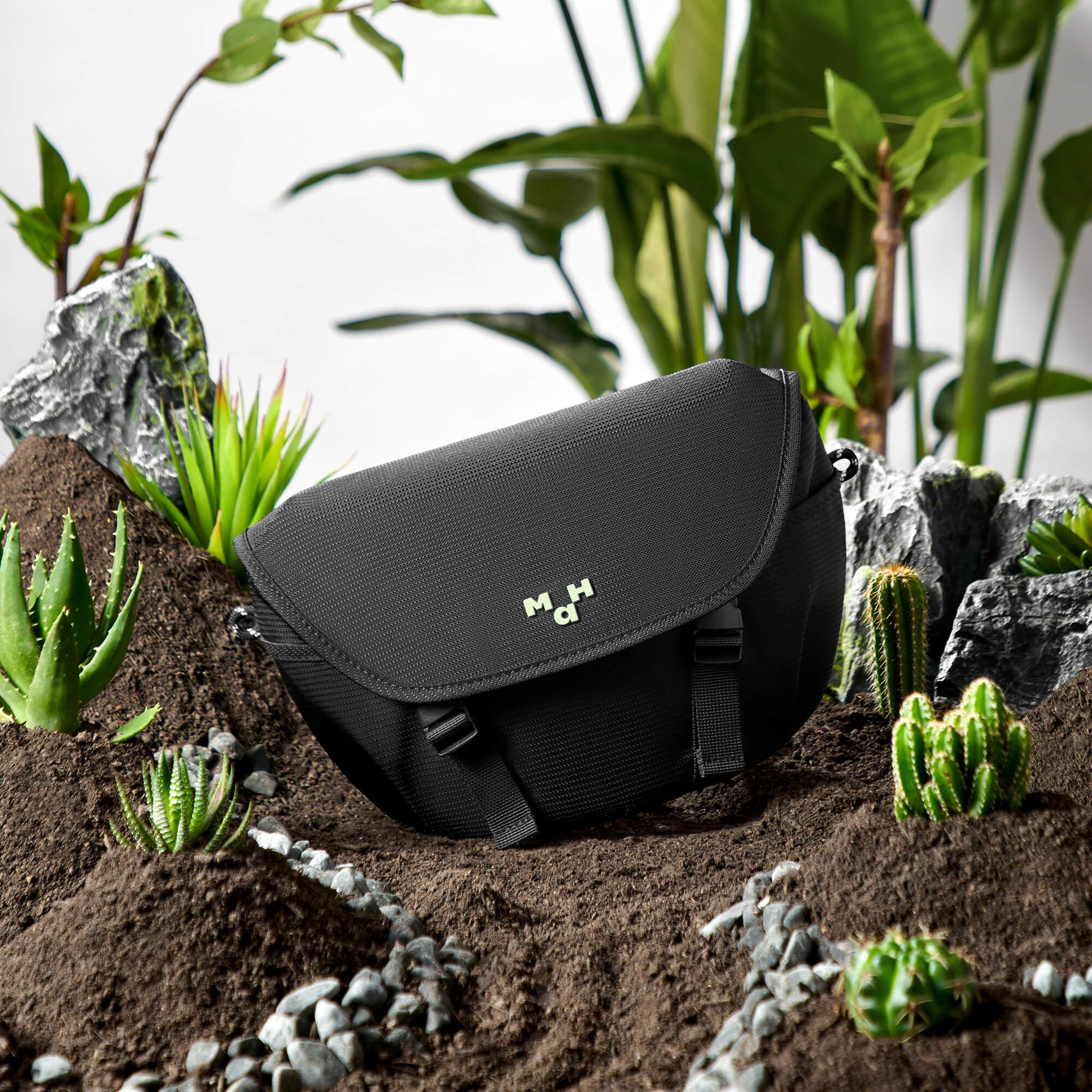 Black sling bag with green logo, styled in desert scene with cacti and succulents for modern lifestyle