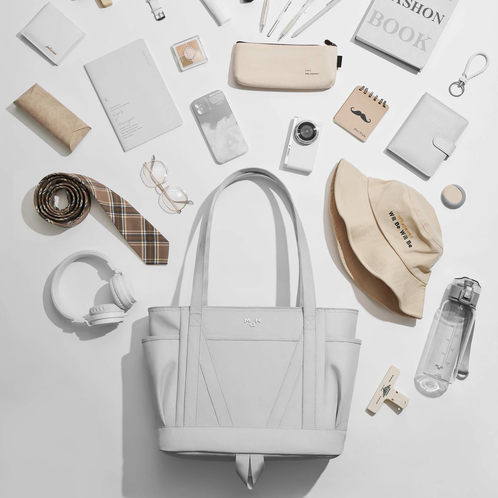 Light gray tote bag with accessories: hat, wallet, headphones, water bottle, and notebook on white background