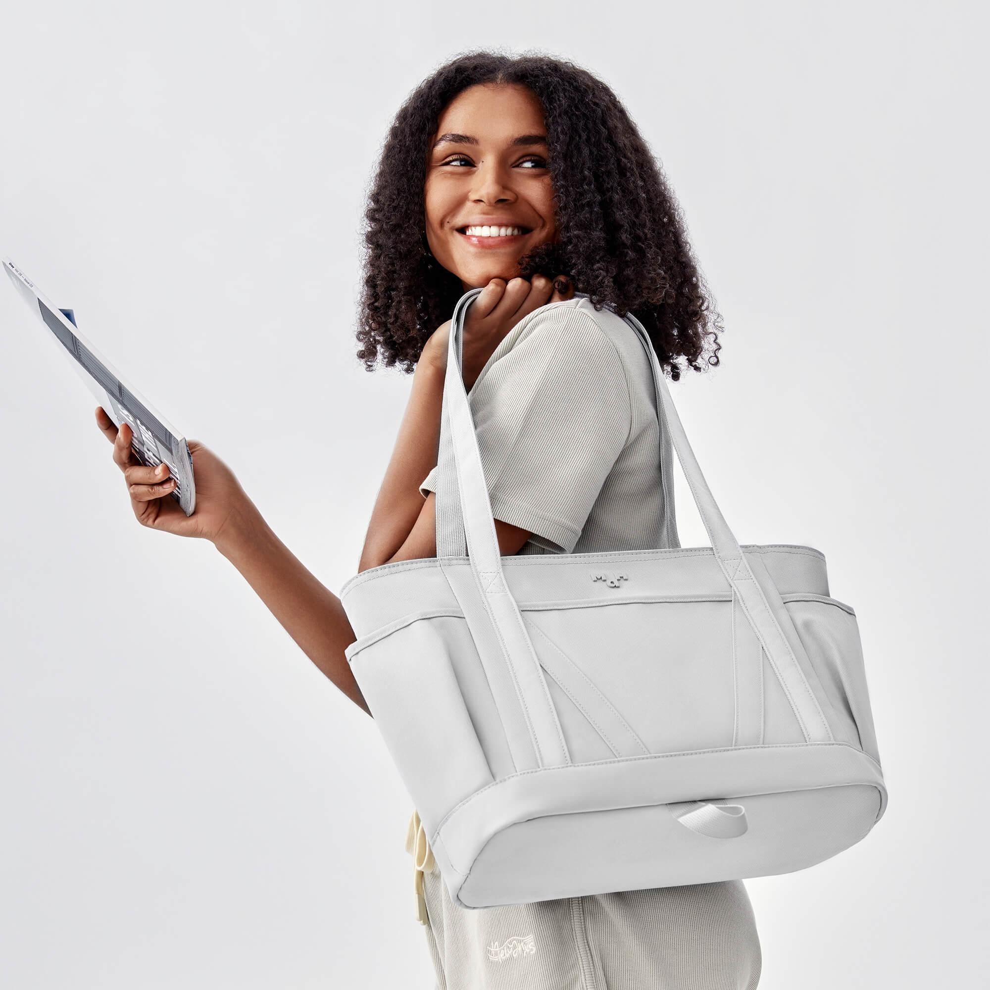 Stylish light gray tote bag with shoulder straps, held by smiling woman in casual outfit, ideal for daily use or travel.