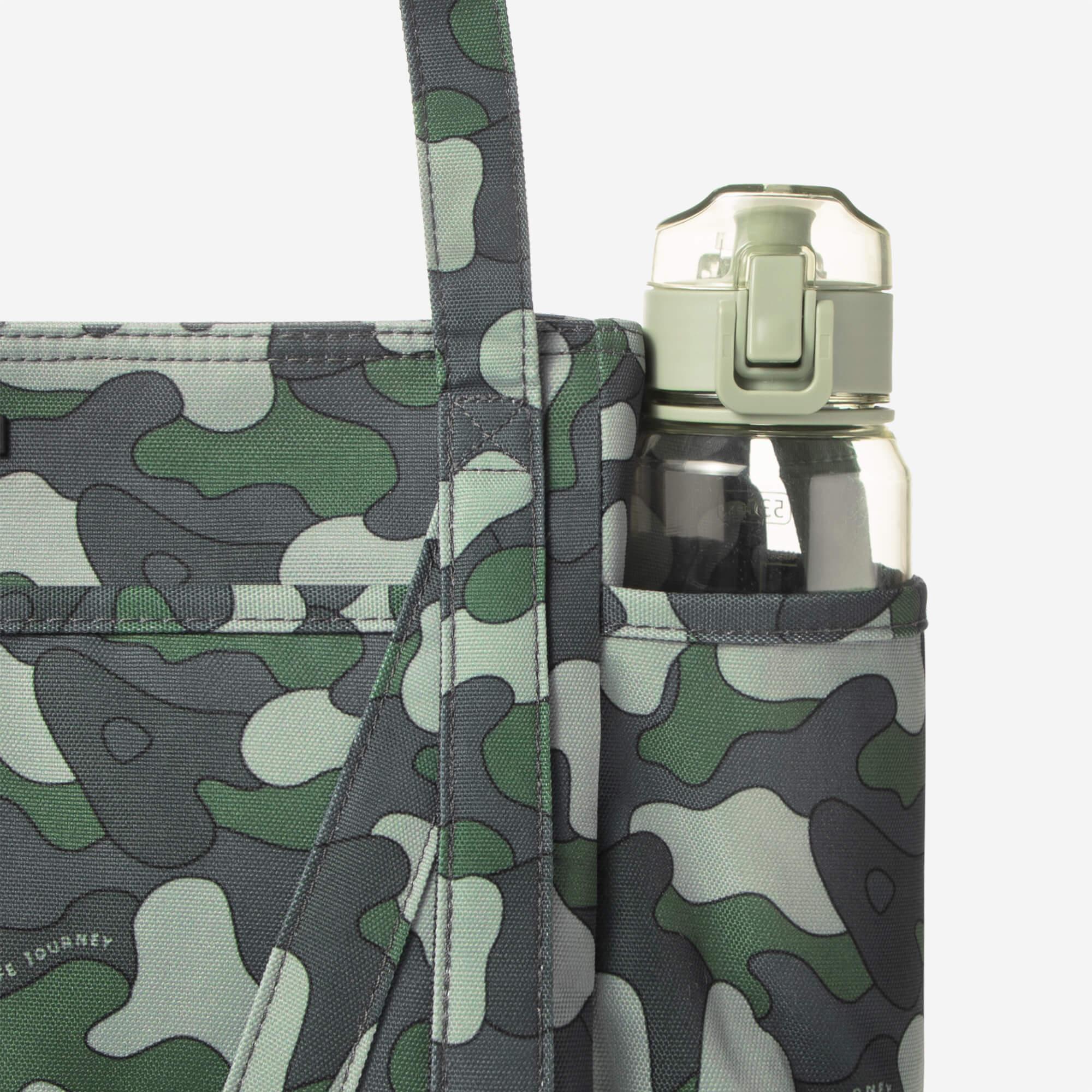 Camouflage tote bag with side water bottle pocket, holding clear reusable bottle, ideal for travel and daily use.