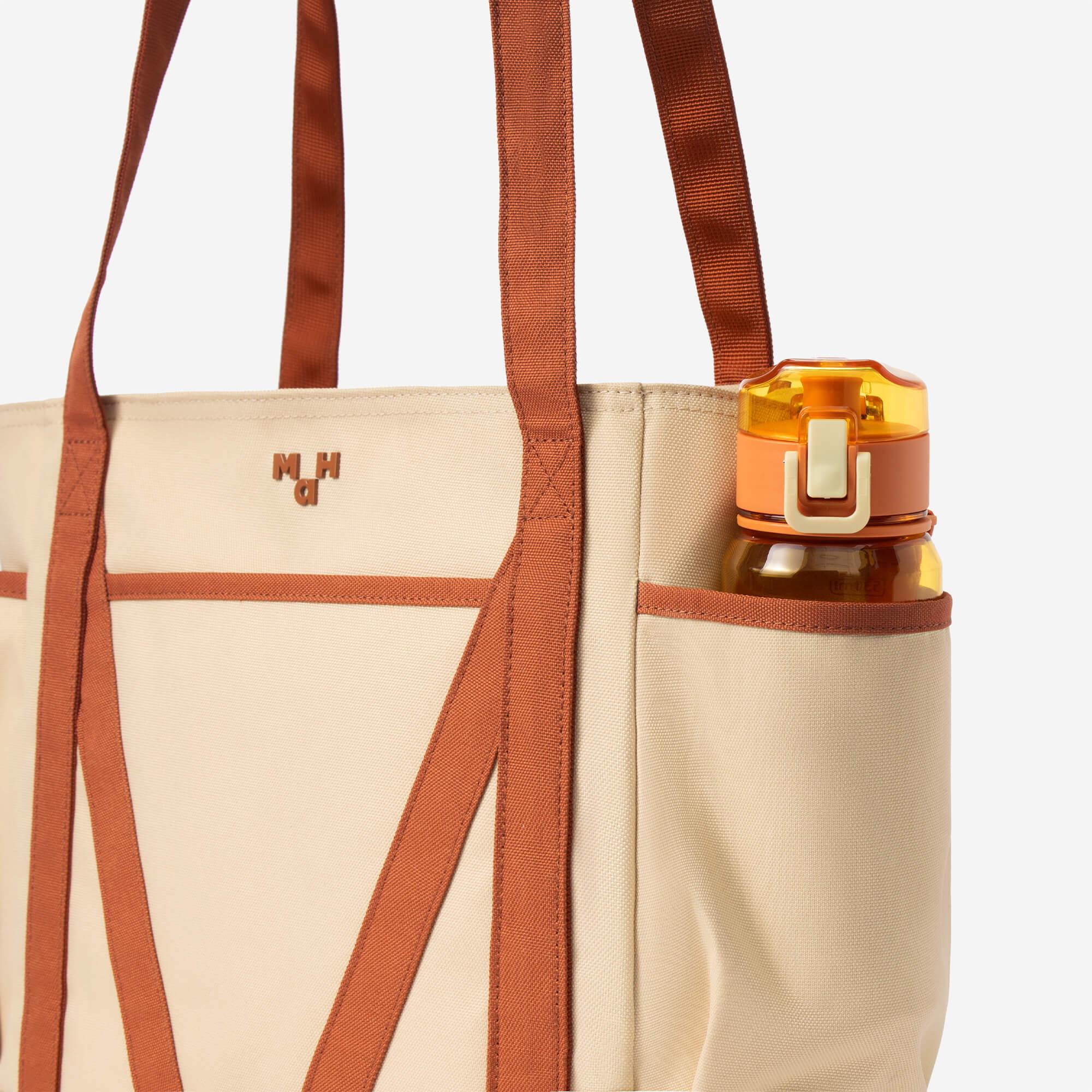 Beige canvas tote bag with rust straps, side pocket holding water bottle, and MH logo detail