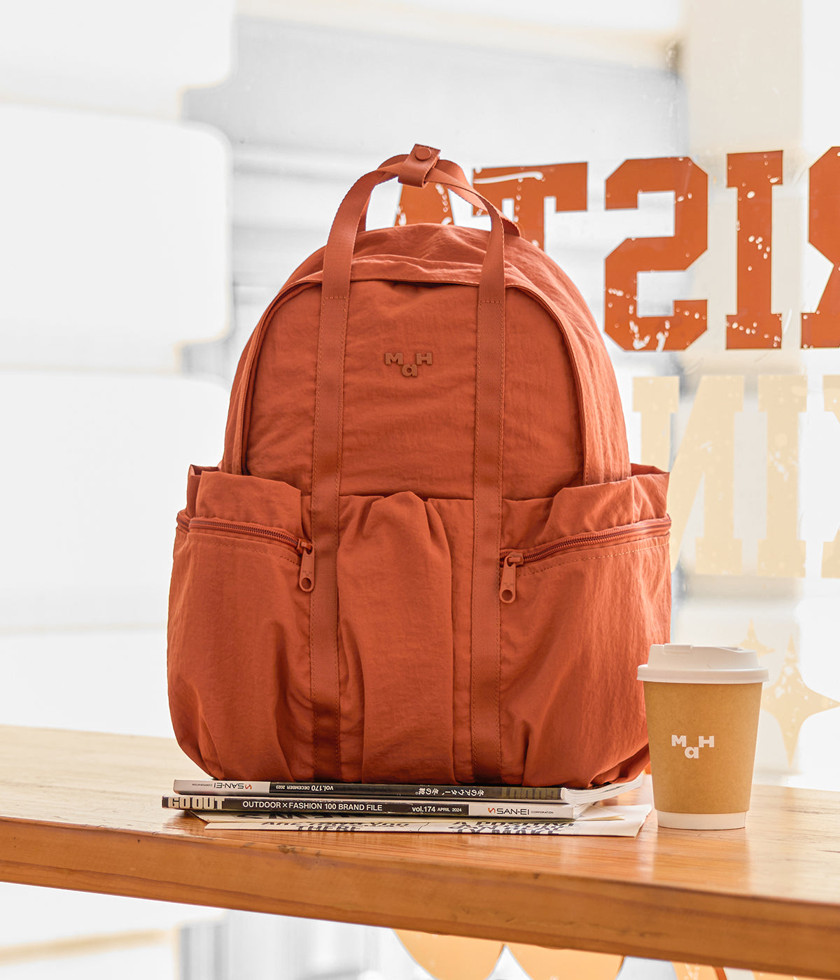 Rust-colored backpack with front pockets on wooden table beside coffee cup and magazines