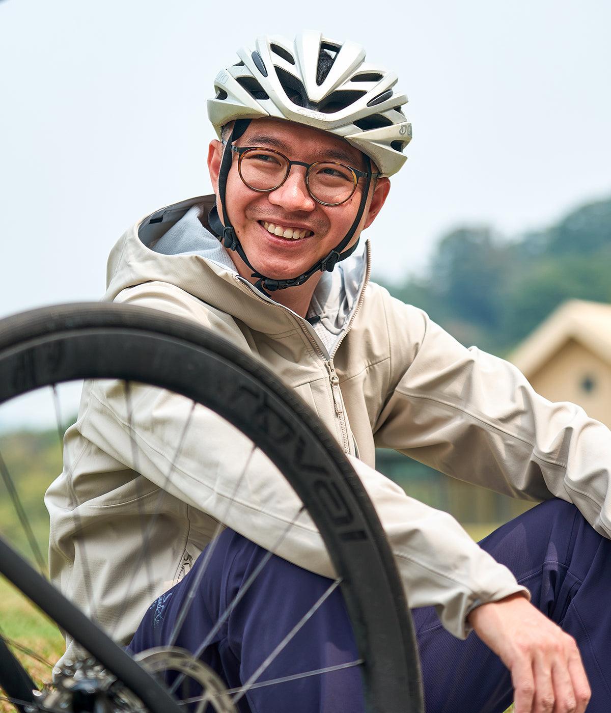 Cyclist in beige jacket and helmet smiling beside bike wheel outdoors