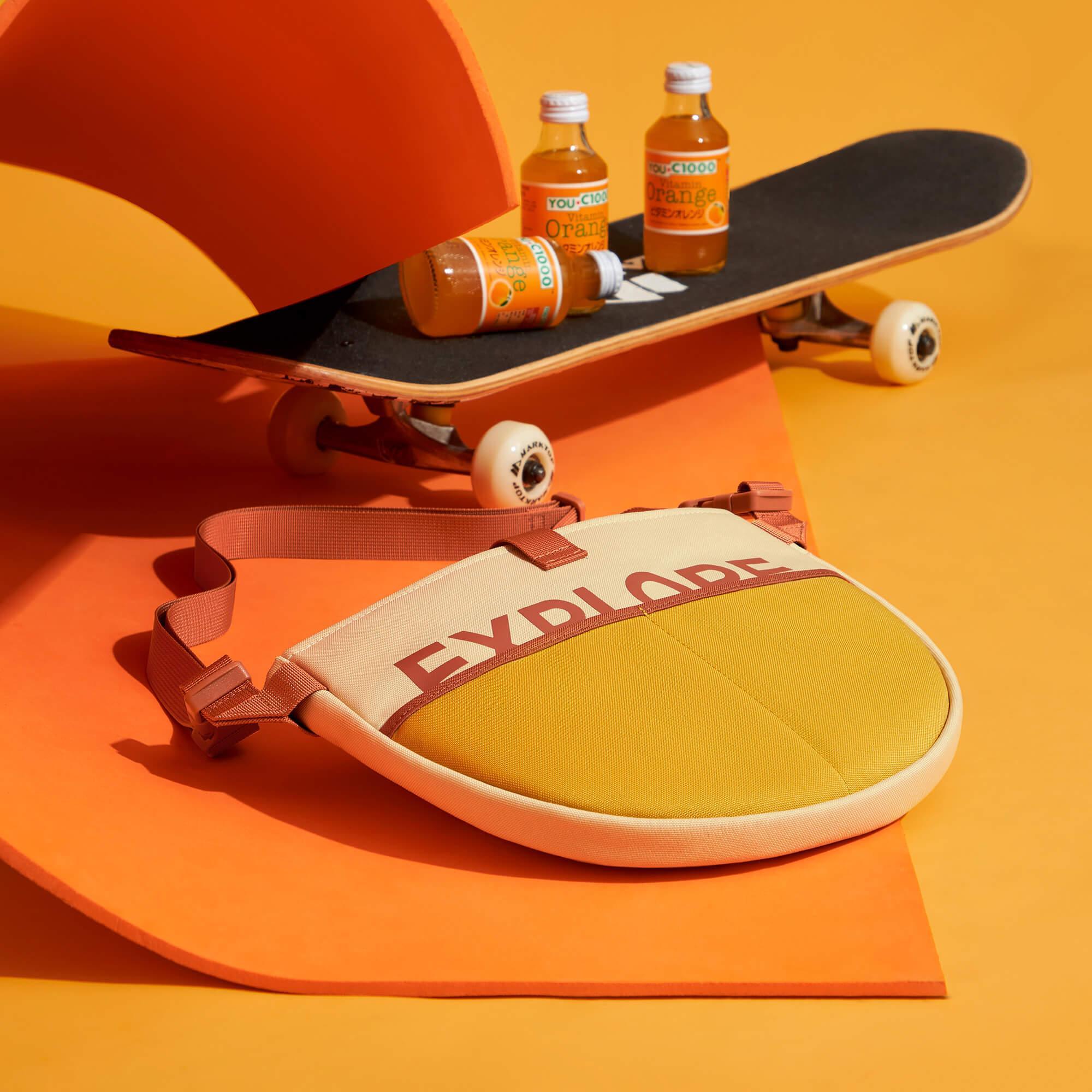 Stylish round crossbody bag with 'EXPLODE' text, paired with skateboard and orange drinks on vibrant orange background