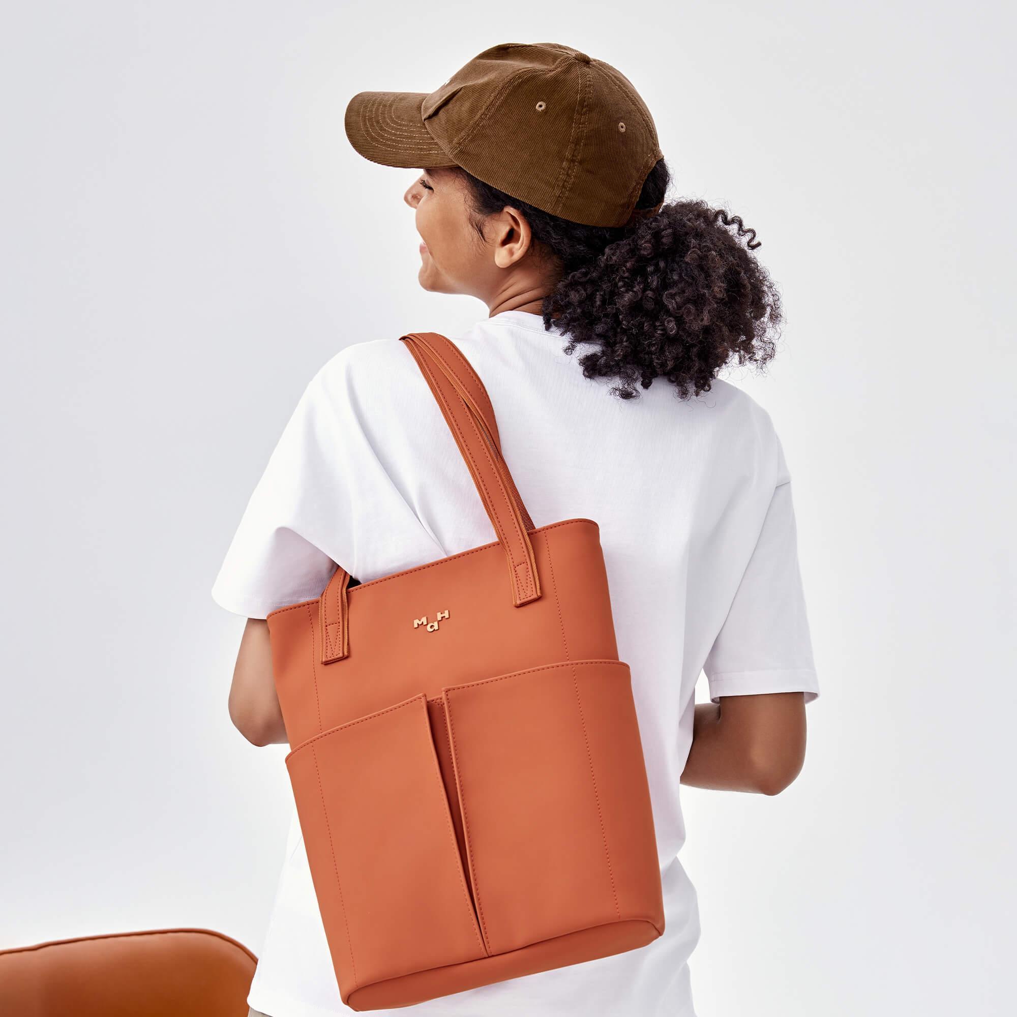 Woman wearing white tee and brown cap carrying orange tote bag with front pockets