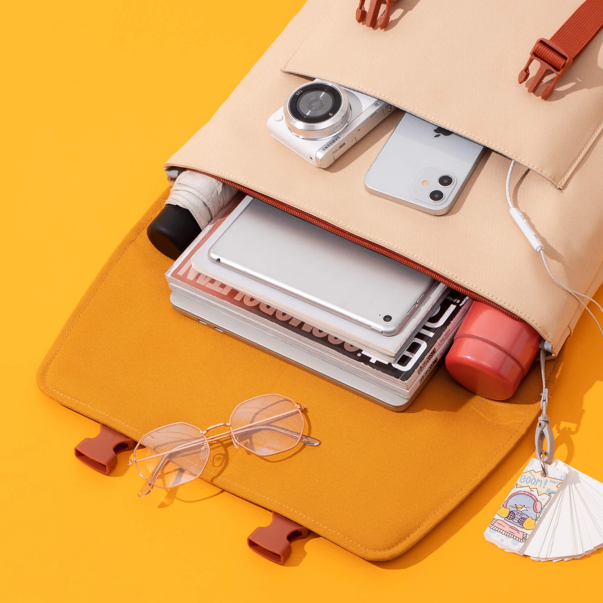 Beige backpack with orange straps, holding camera, phone, tablet, magazines, and glasses on yellow background