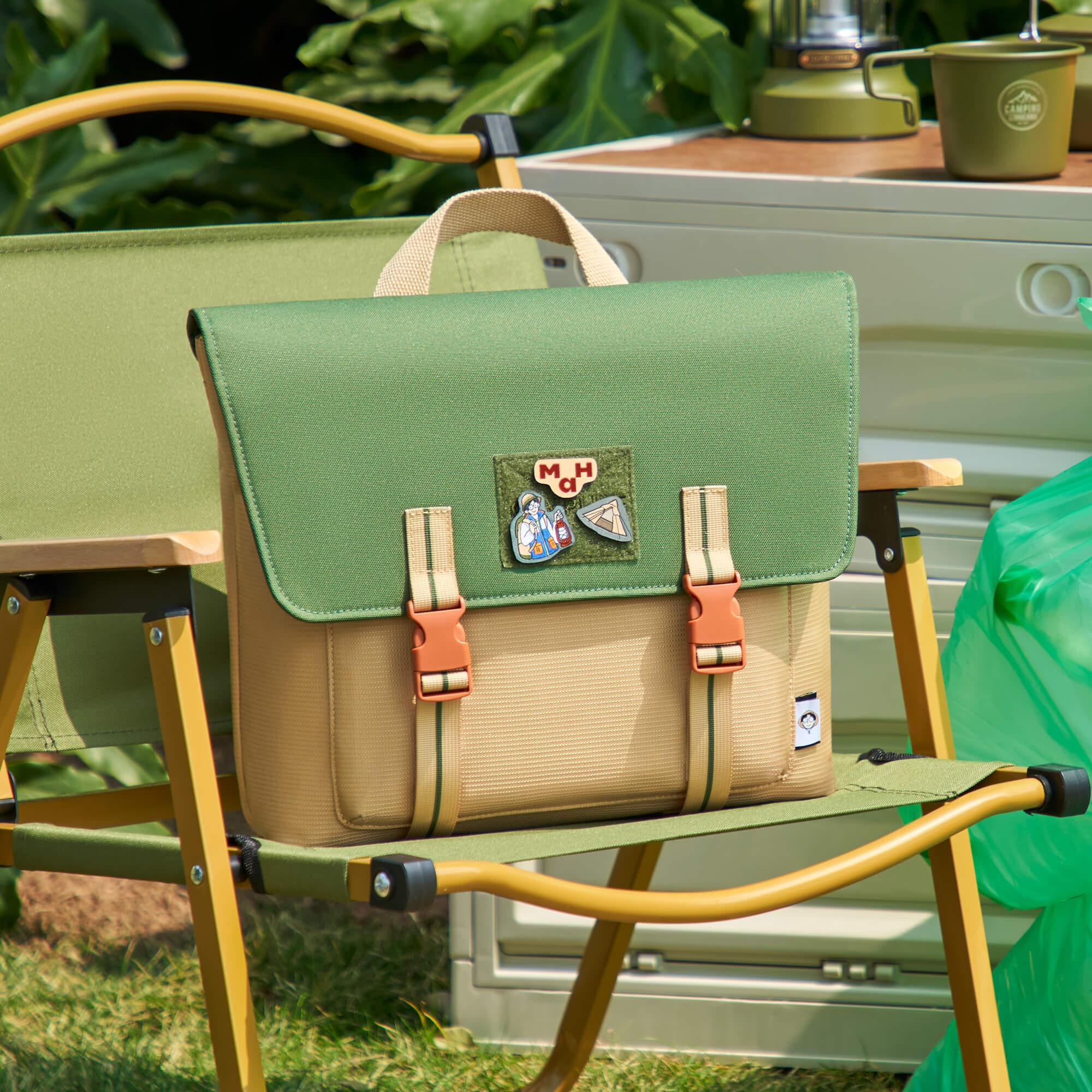 Green and beige canvas satchel bag with patches, resting on a camping chair outdoors