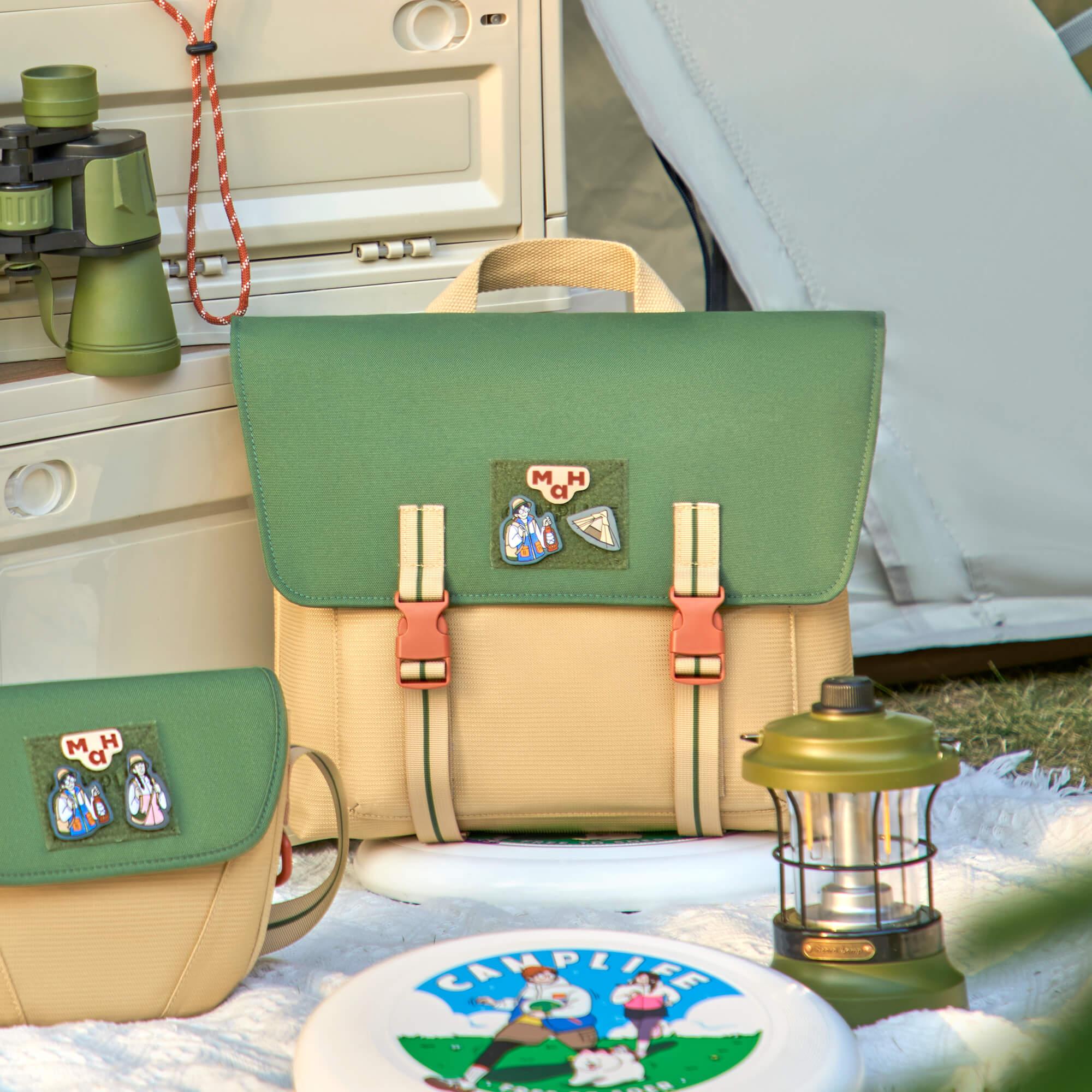 Green and beige camping satchel with patches, binoculars, lantern, and frisbee on picnic blanket near tent