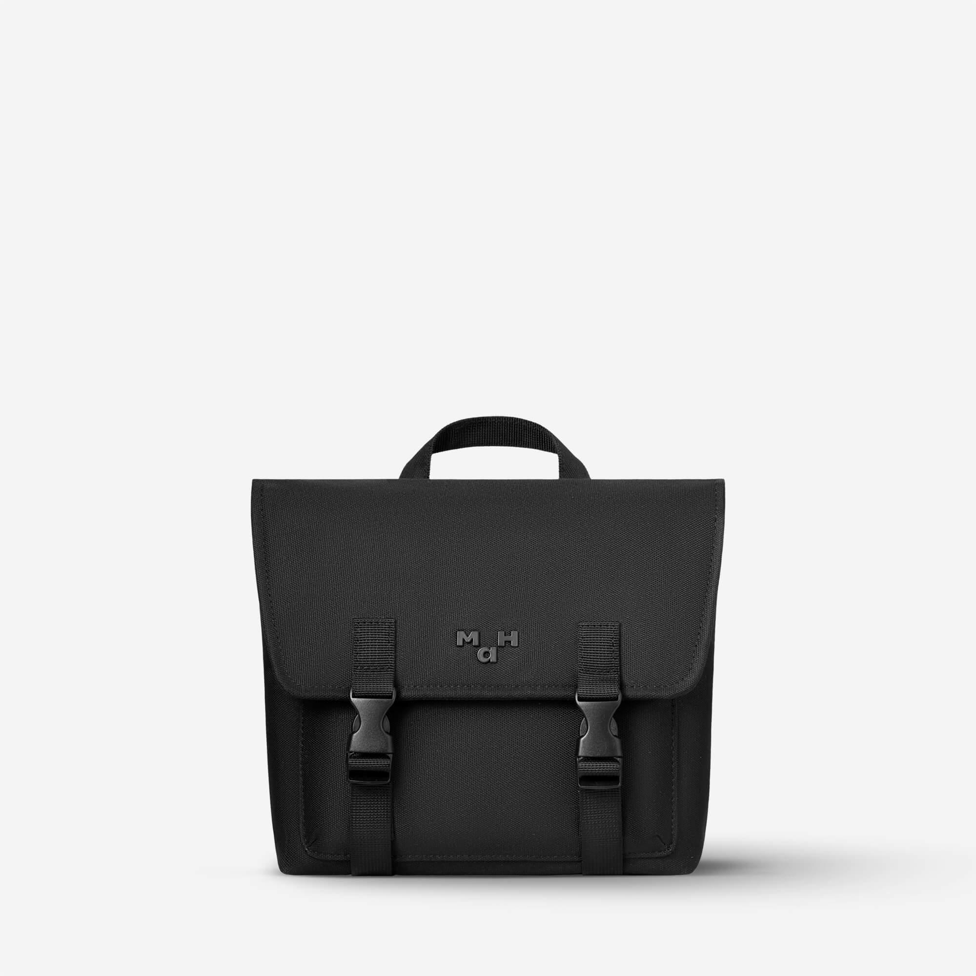 Black minimalist satchel bag with front flap, buckles, and top handle, ideal for everyday carry and urban style.