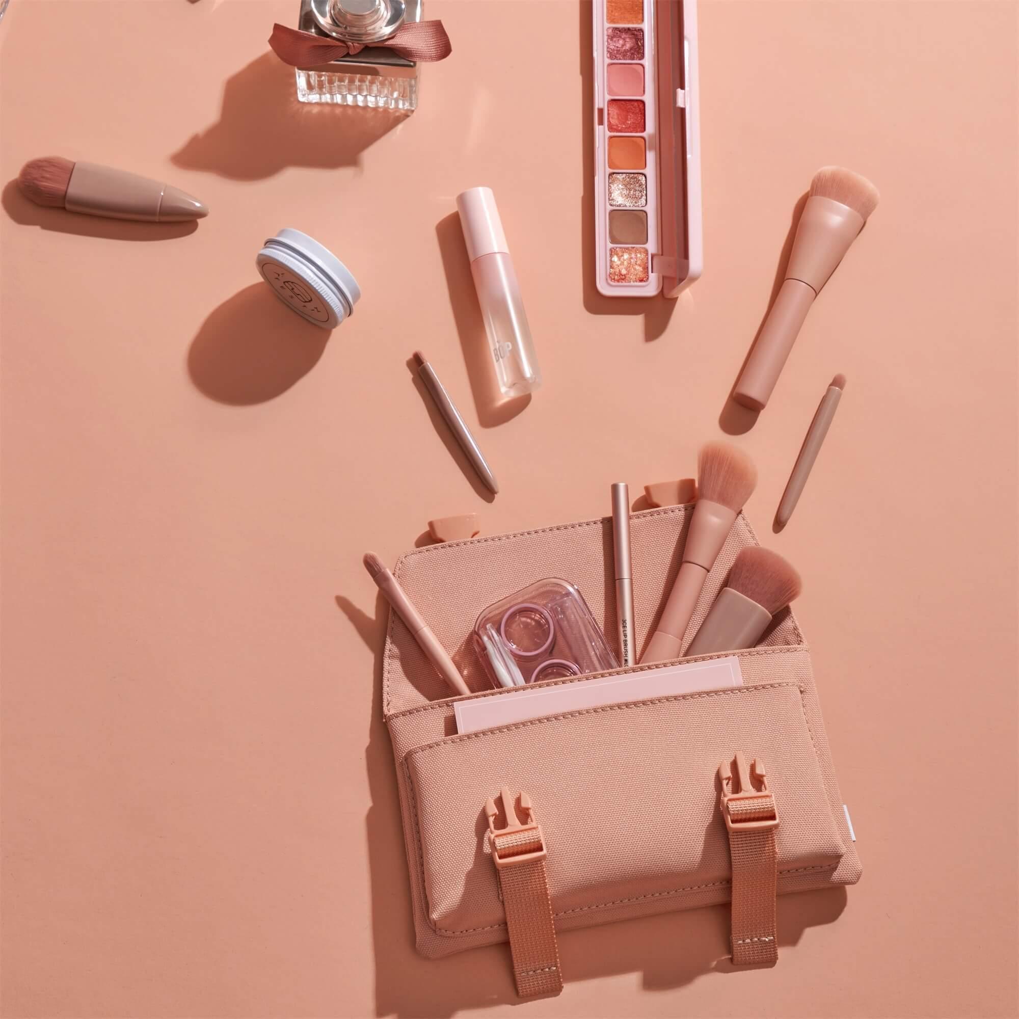 Pink makeup bag with brushes, eyeshadow palette, and cosmetics on soft peach background