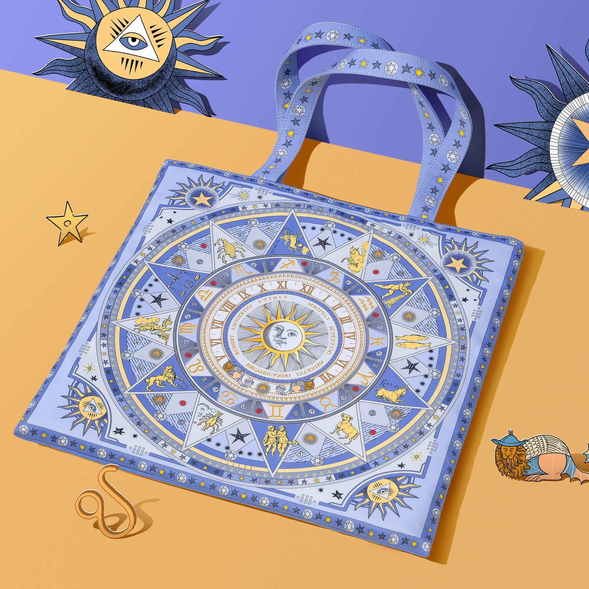 Blue tote bag with zodiac and celestial print, featuring sun, stars, and astrological symbols on a purple and gold background.
