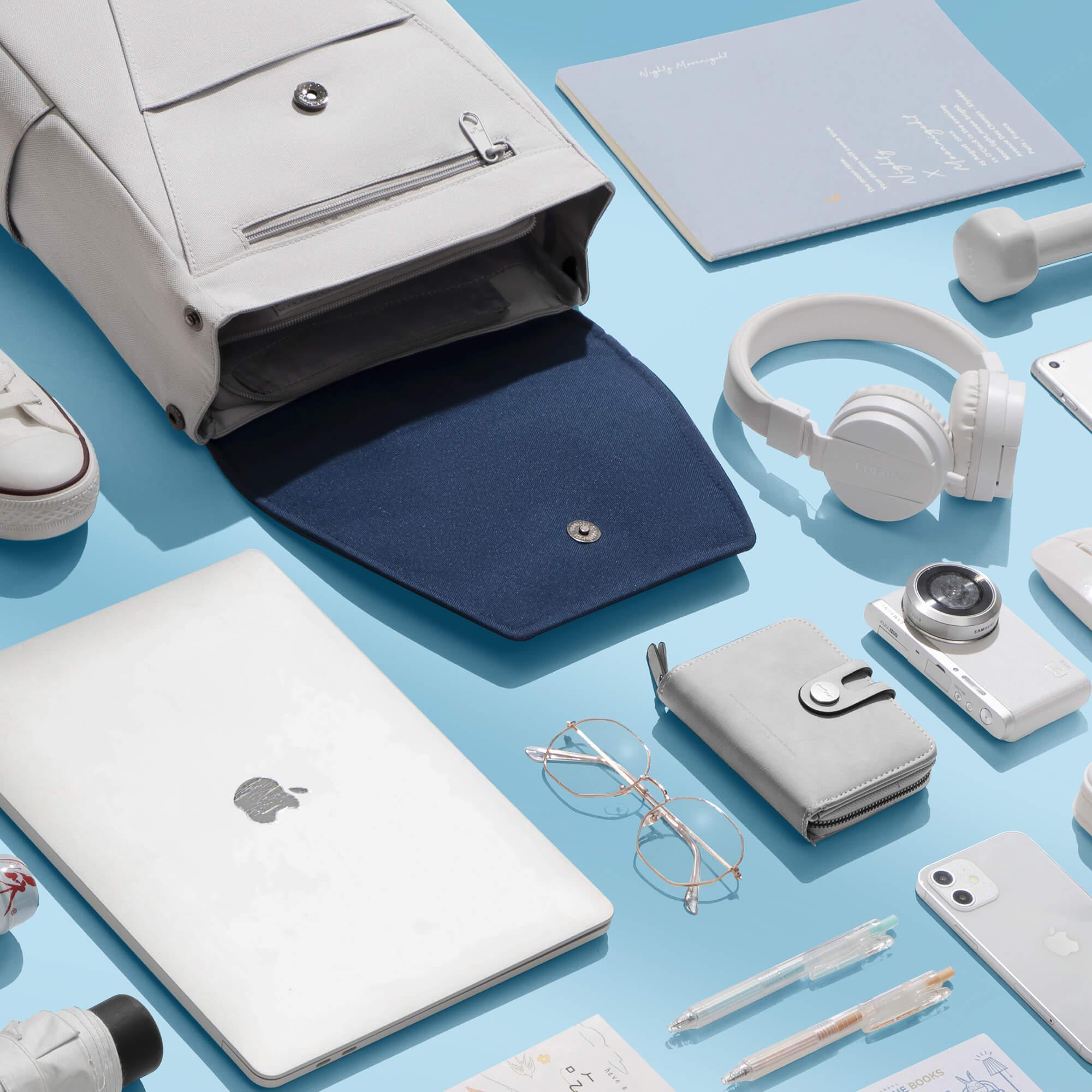 Light gray backpack with navy interior, surrounded by laptop, headphones, wallet, and daily essentials on blue background