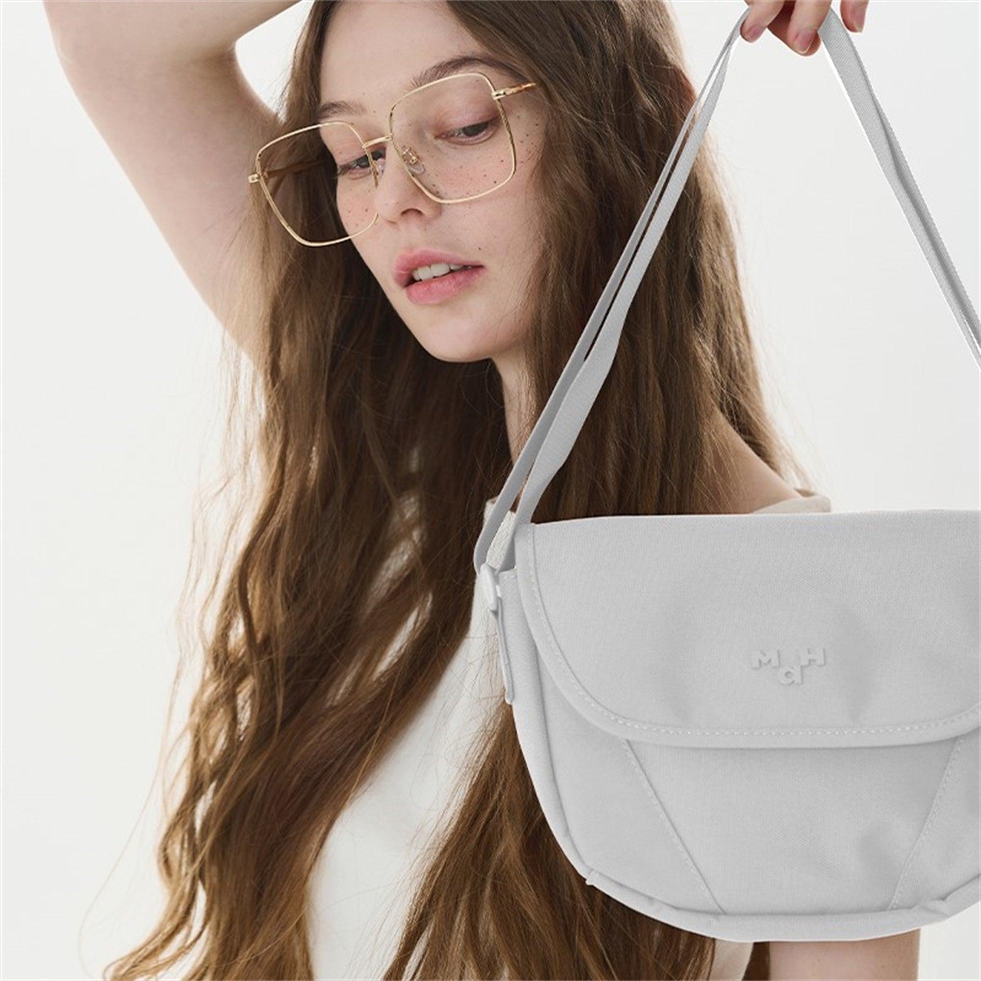 Woman holding light gray crossbody bag with adjustable strap, minimalist design, and subtle logo detail.