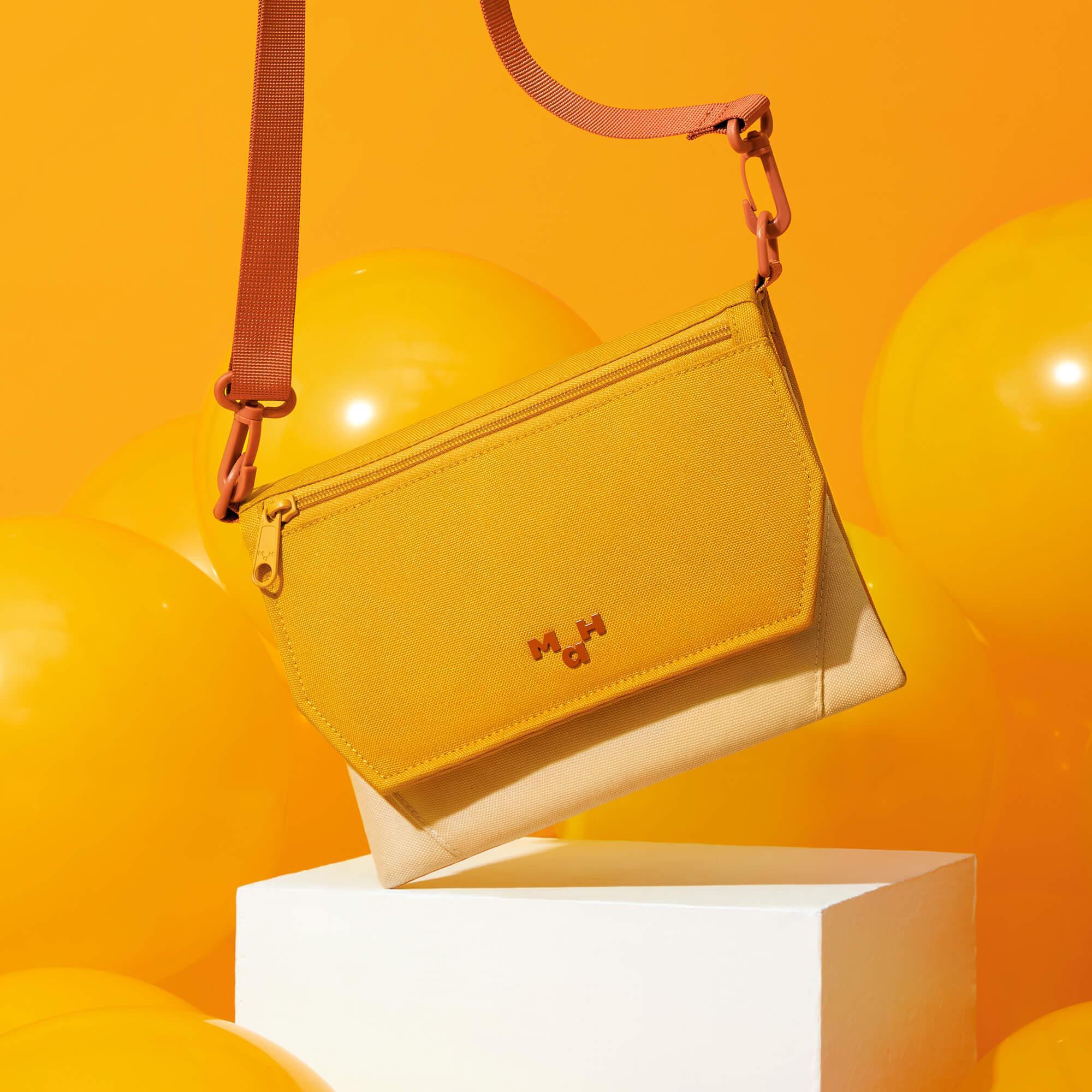 Mustard yellow crossbody bag with adjustable strap, modern design, displayed on white pedestal against vibrant orange background