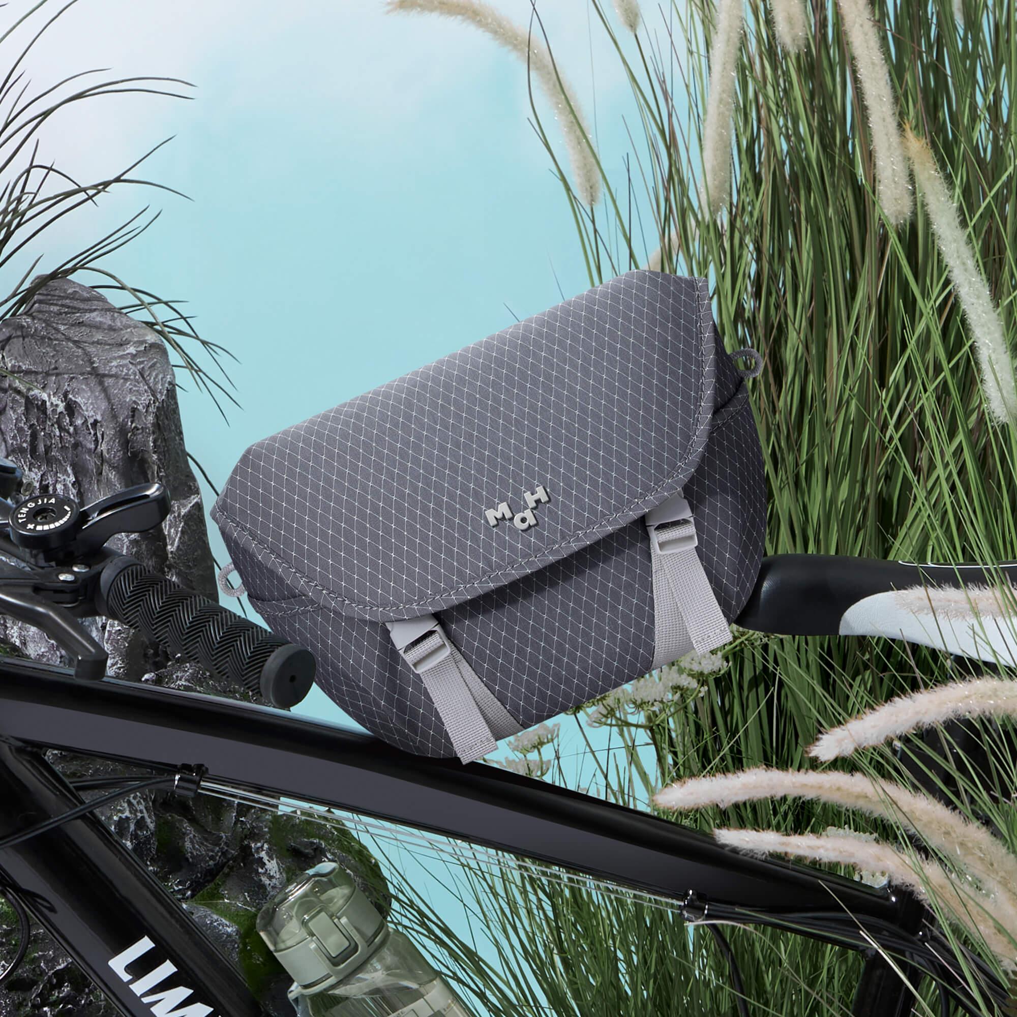 Gray diamond-pattern bike bag with MaH logo, attached to bicycle handlebars near grassy field