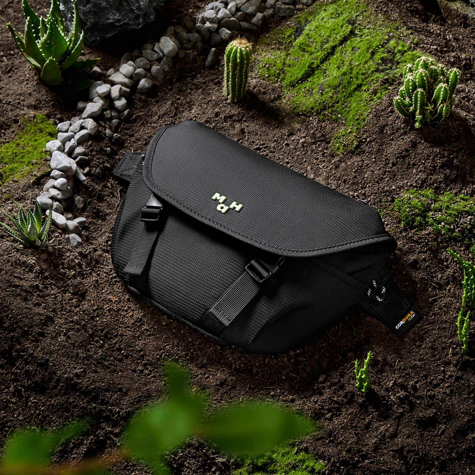 Black durable sling bag with MH logo, set on earthy ground with succulents and moss