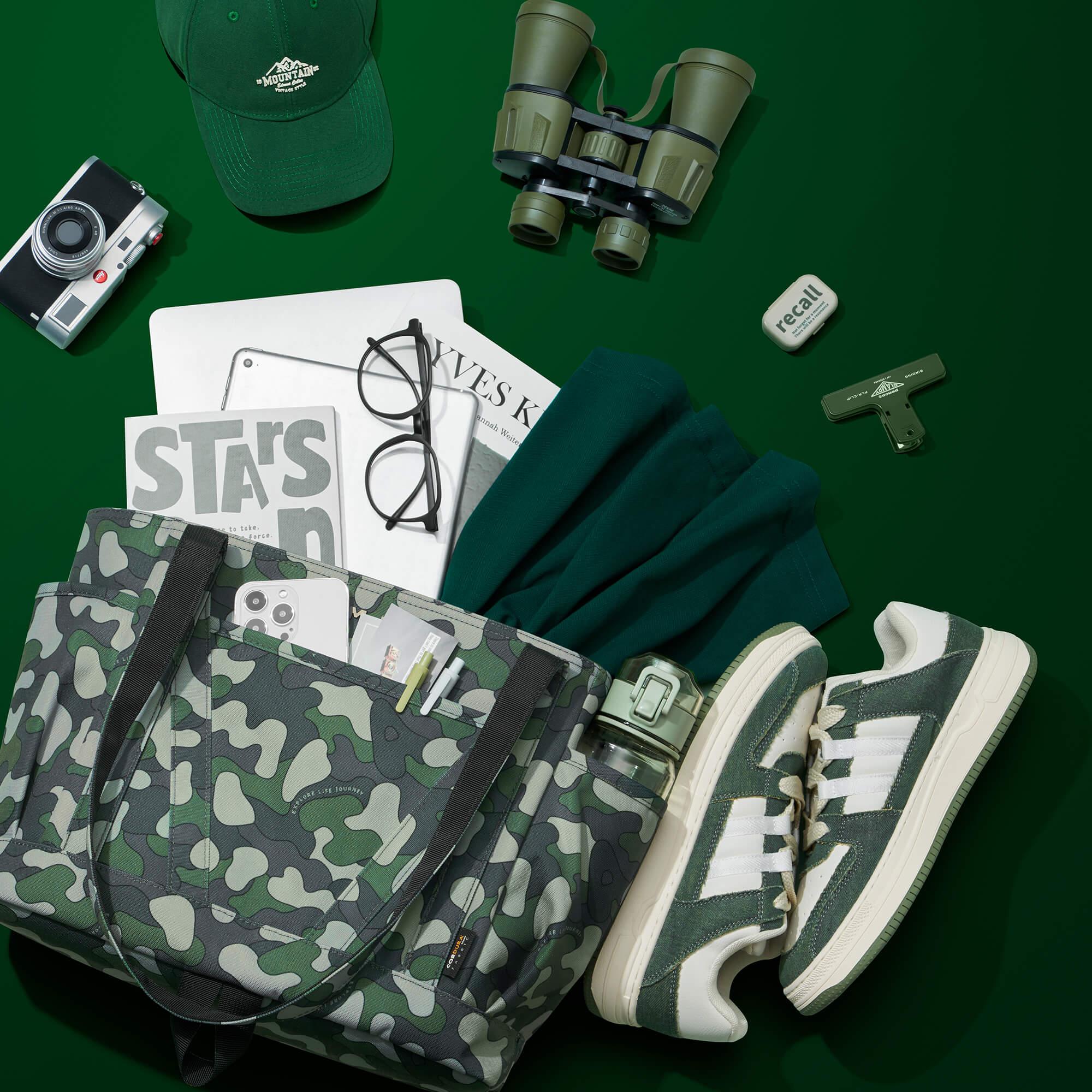Camouflage tote bag with travel essentials: green sneakers, cap, binoculars, camera, and accessories on green background