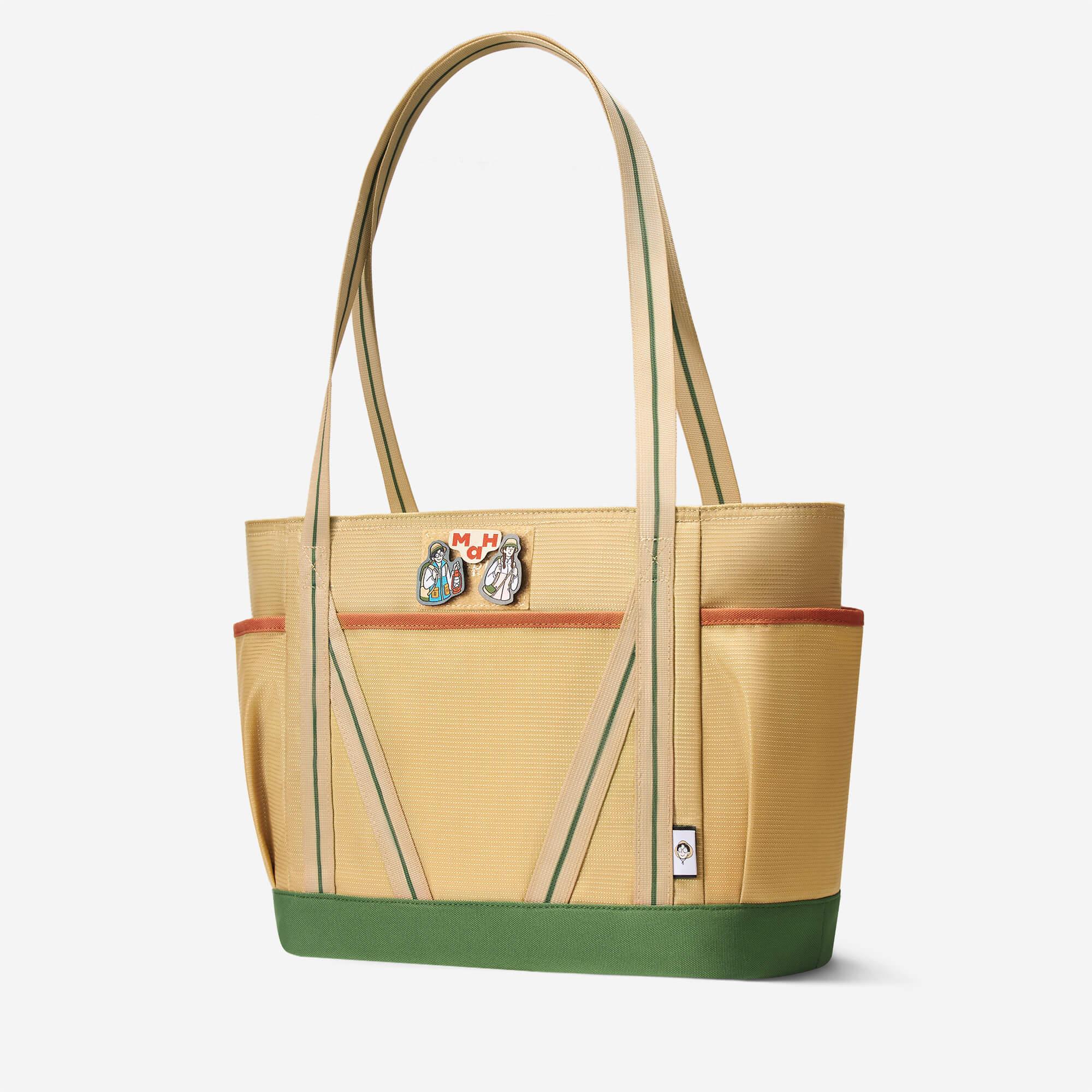 Beige tote bag with green stripes, front pockets, and character pins, ideal for daily use or travel.