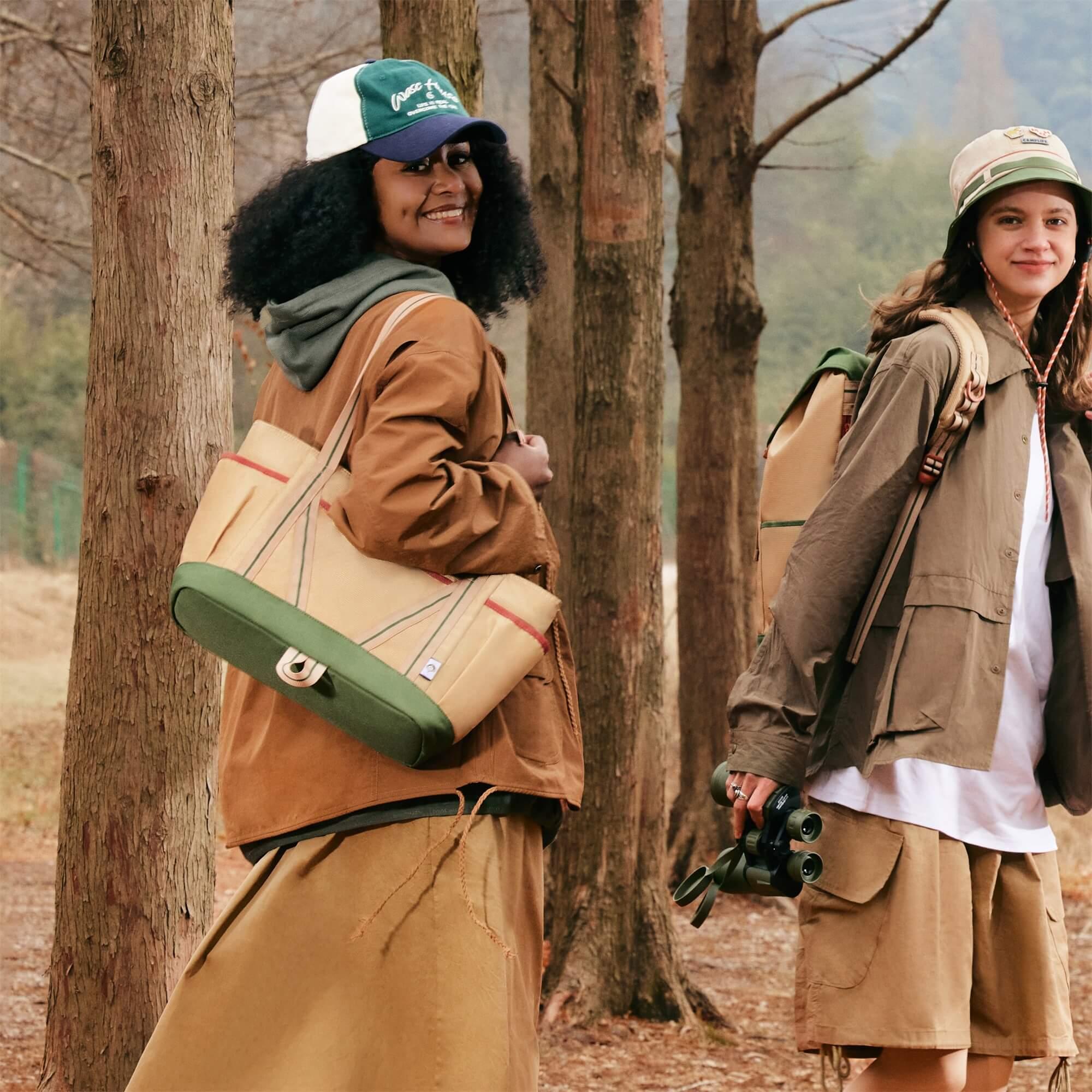 Two women in outdoor gear with canvas tote bags and binoculars in a forest setting